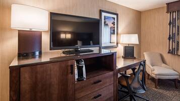 A hotel room with a wooden desk, a flat-screen TV, a minibar, a chair, and a framed picture on the wall.