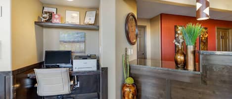 A reception desk with a computer, printer, and framed certificates. There is a vase with green plants and a decorative wall piece.