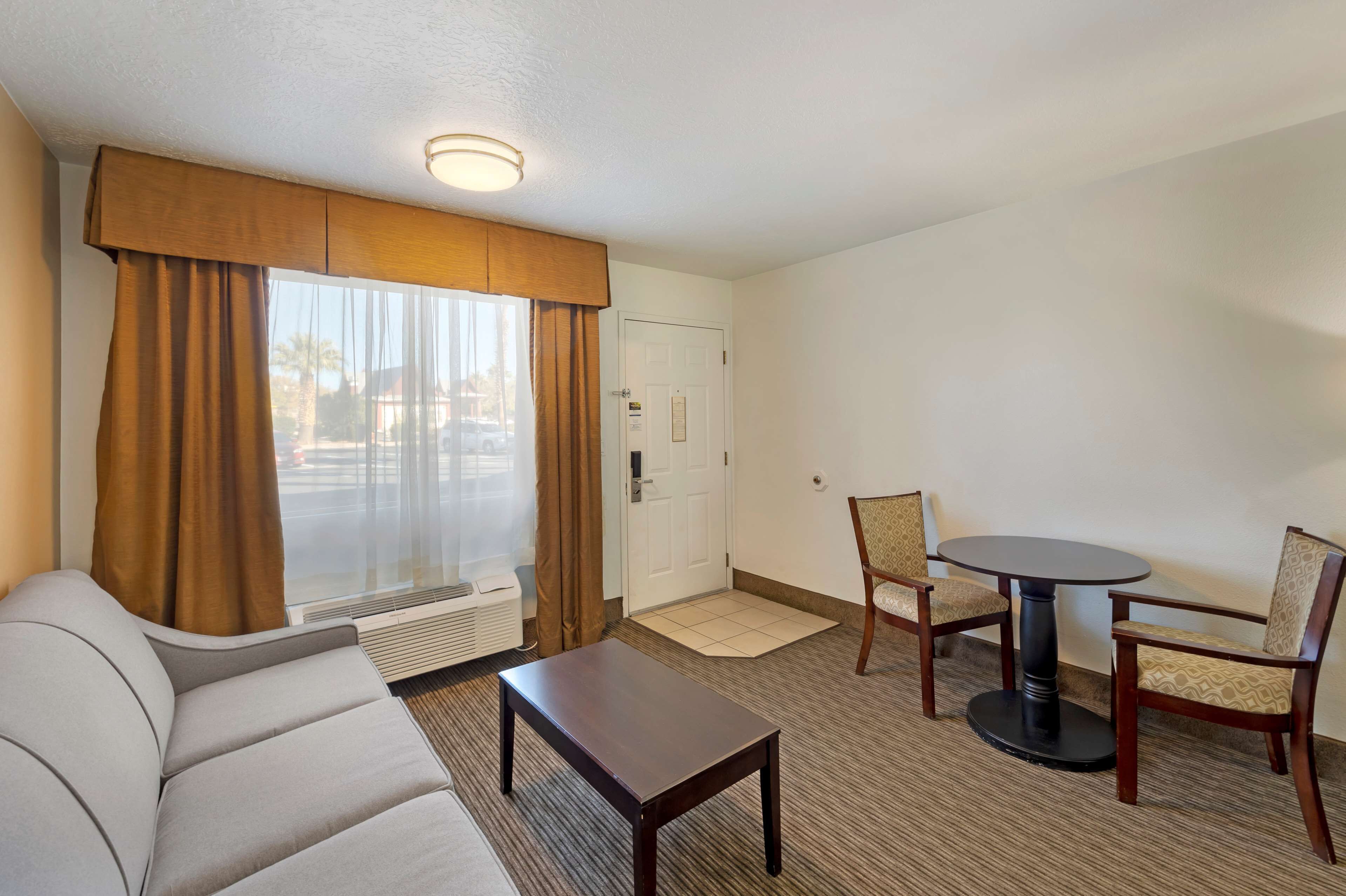 A hotel room with a sofa, a small table, two chairs, a round table, a door, and a window with curtains.