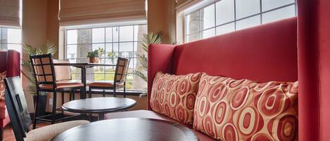 A seating area with a red sofa, two chairs, and a round table. There are potted plants and a view through large windows.
