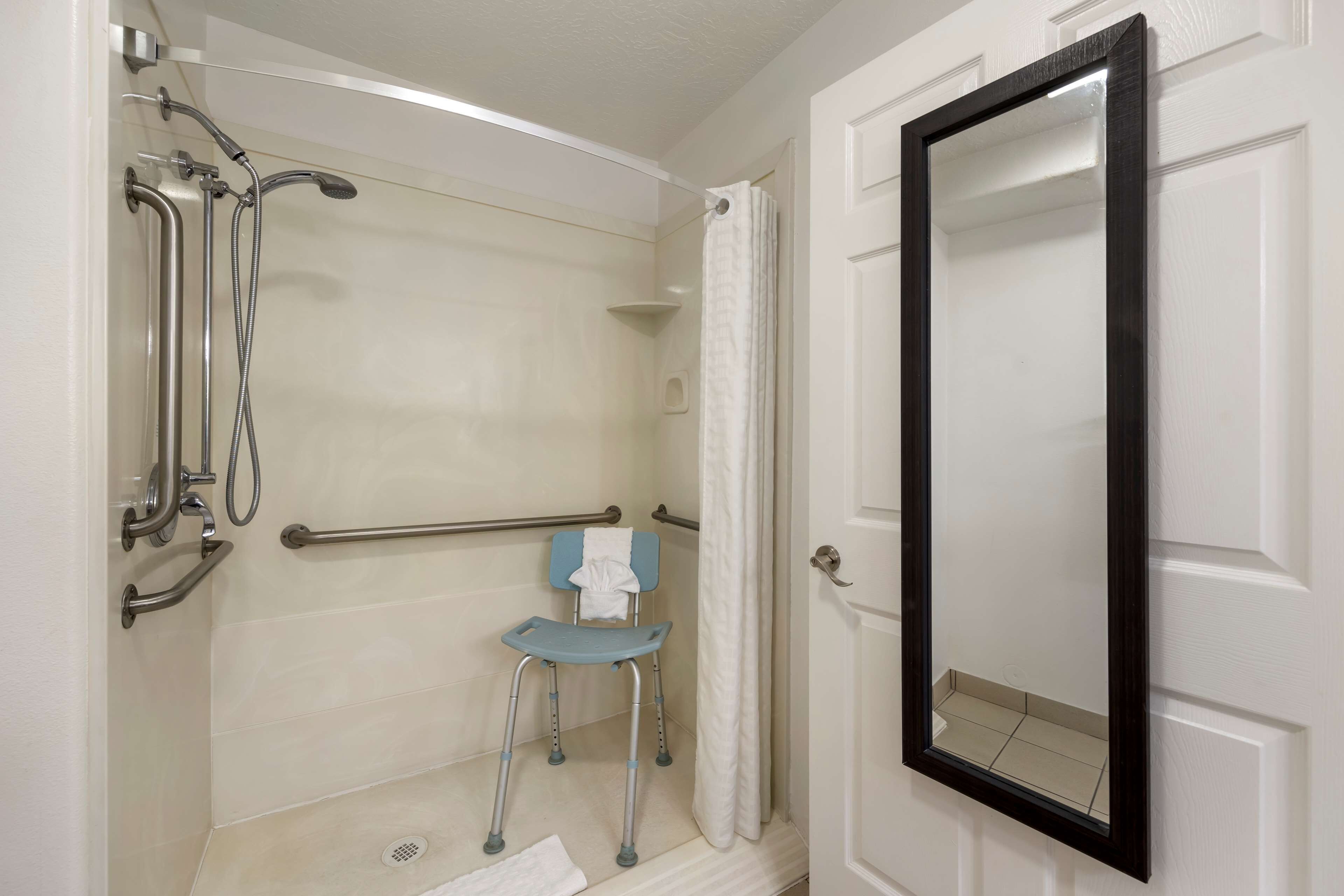 Une salle de bain équipée d’une douche, de barres d’appui, d’un siège accessible aux personnes en fauteuil roulant et d’une porte avec un miroir.