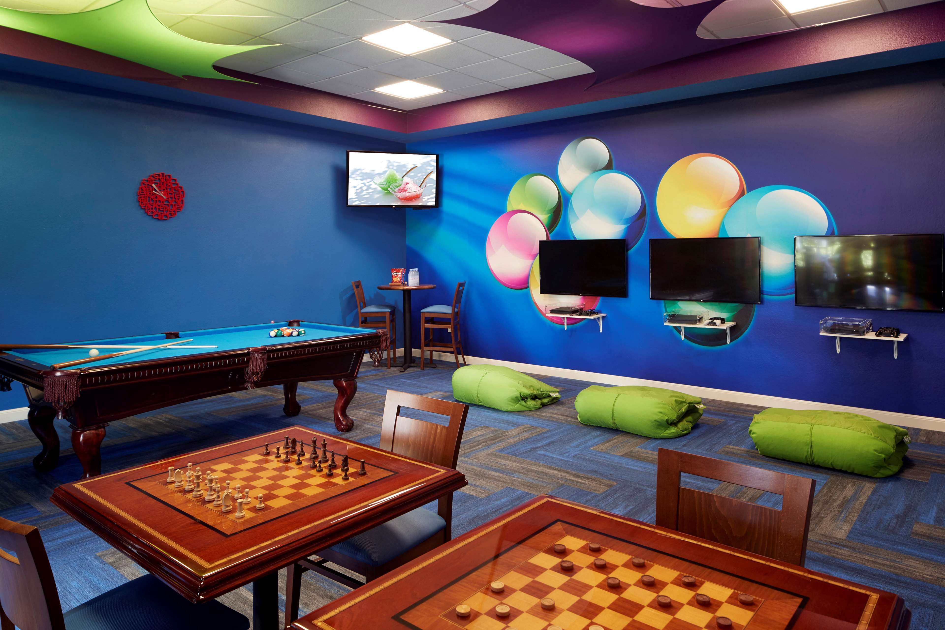 A room with a pool table, a billiards table, and a chess table. There are green bean bags and a TV mounted on the wall.