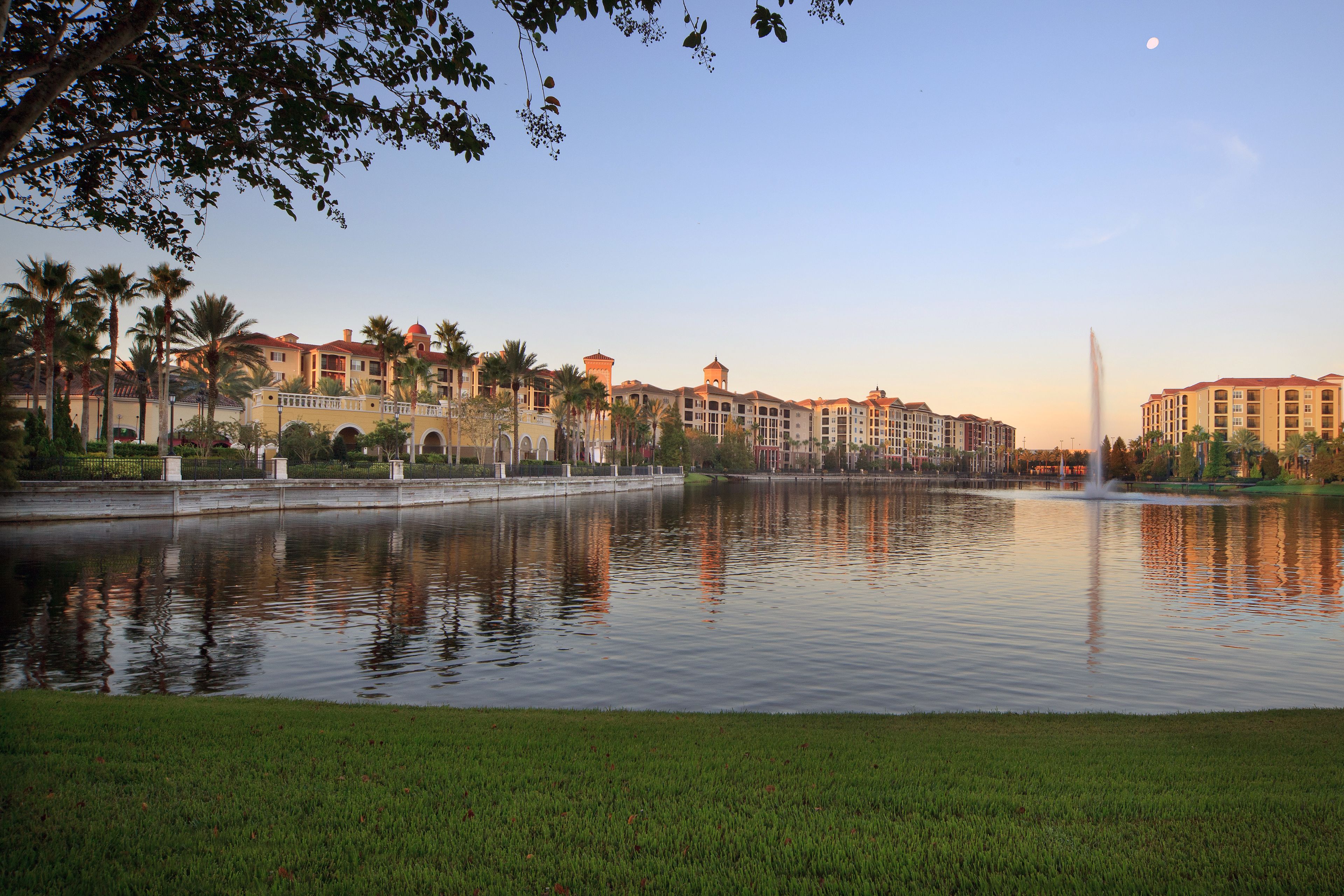 A large body of water with a fountain in the center, surrounded by a well-maintained lawn and palm trees. In the background, there are multi-story buildings with a uniform architectural style.