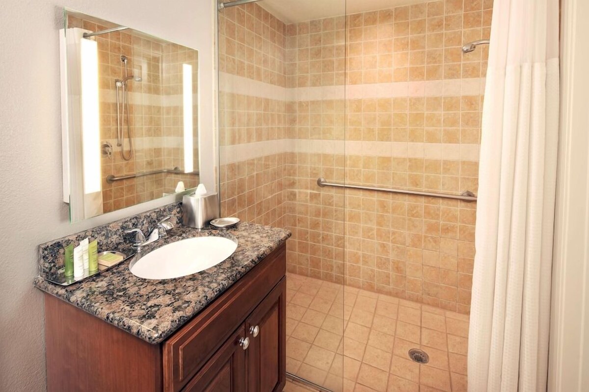 A bathroom with a granite countertop sink, a mirror, and a shower with beige tiles.