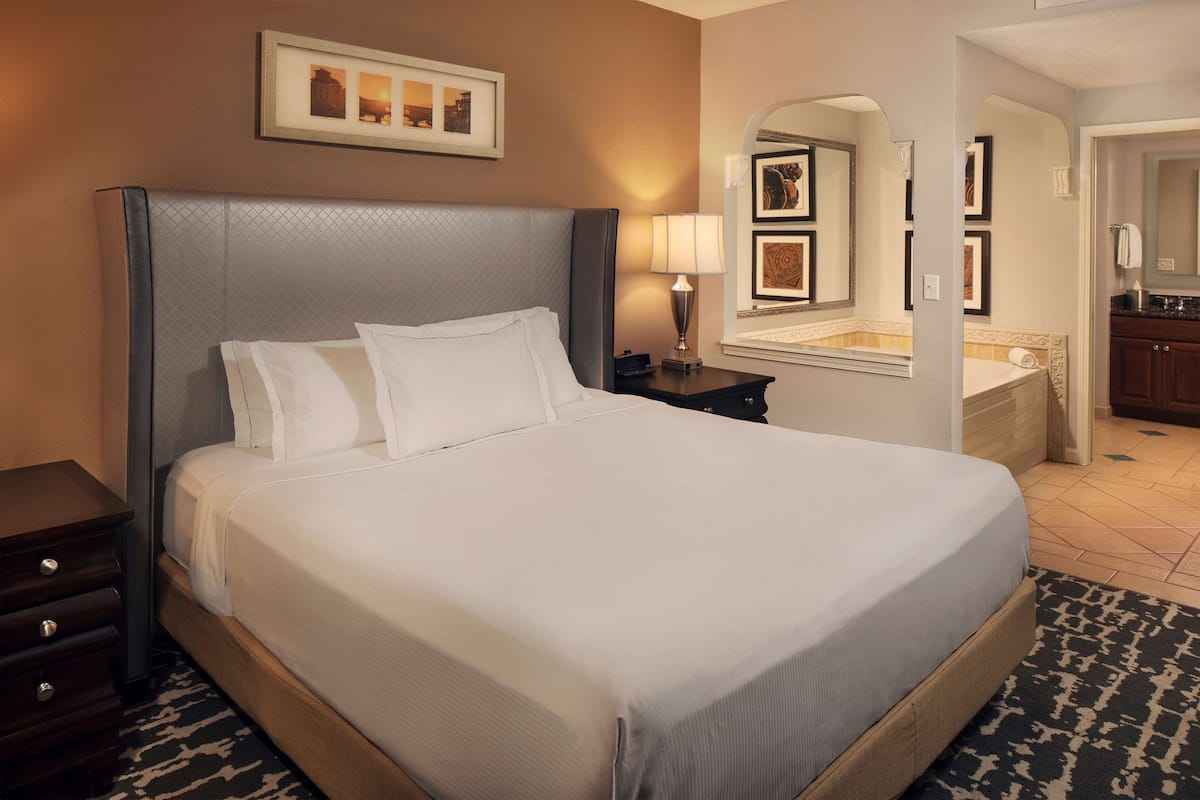 Een hotelkamer met een groot bed, een nachtkastje met een lamp, een lijst met een afbeelding boven het bed en een badkamer zichtbaar door een boog.