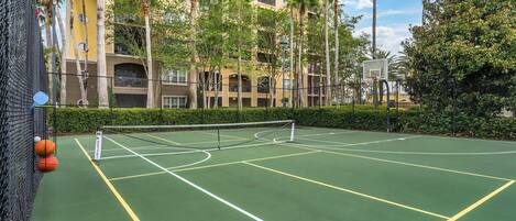Un court de tennis avec un filet, entouré d’une clôture, et situé à côté d’un immeuble doté de balcons et de palmiers.