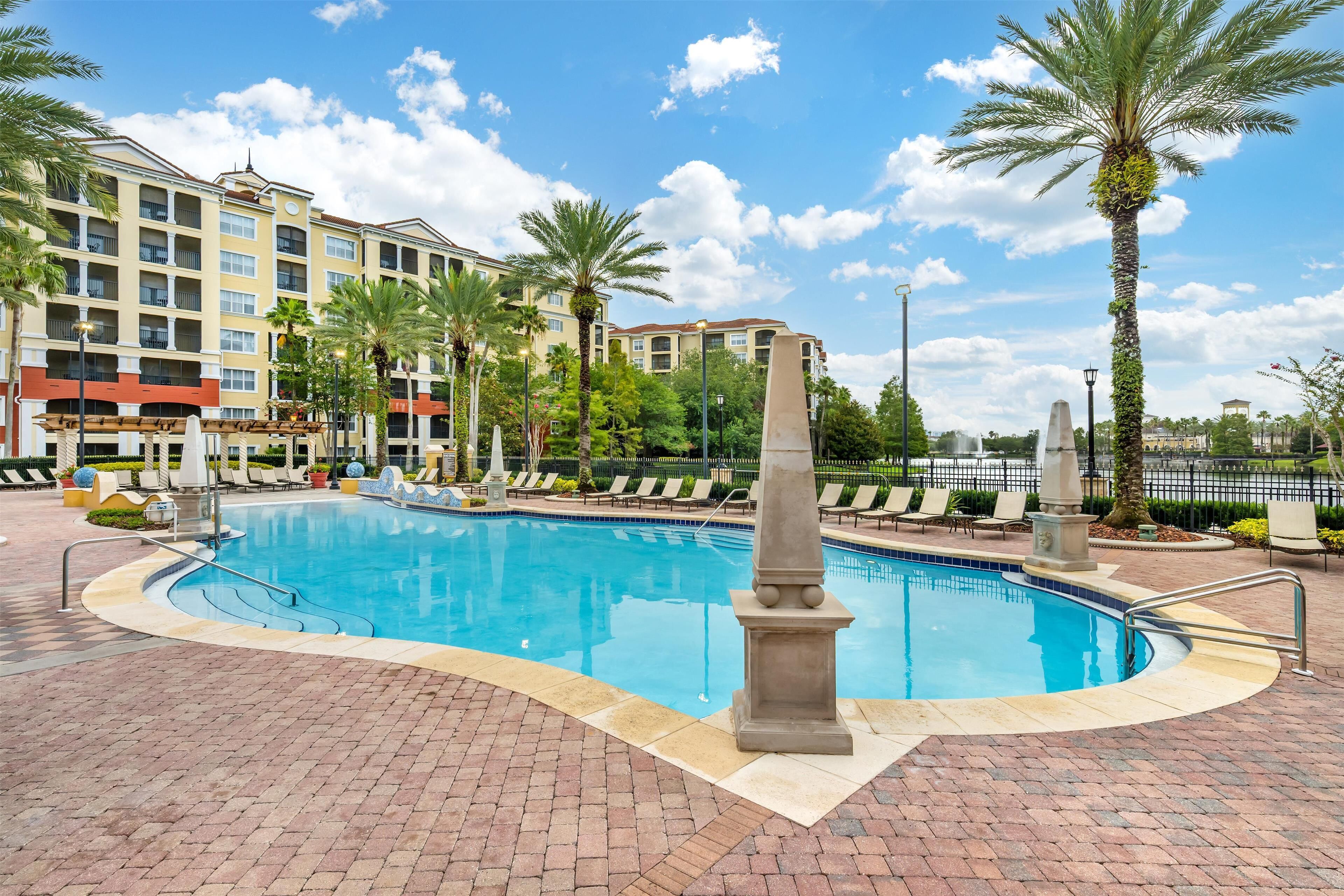A swimming pool with a stone pillar, surrounded by lounge chairs and palm trees, with a hotel building in the background.