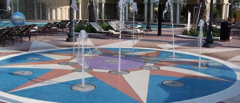 A circular water fountain with multiple jets, surrounded by a tiled area with lounge chairs and palm trees.