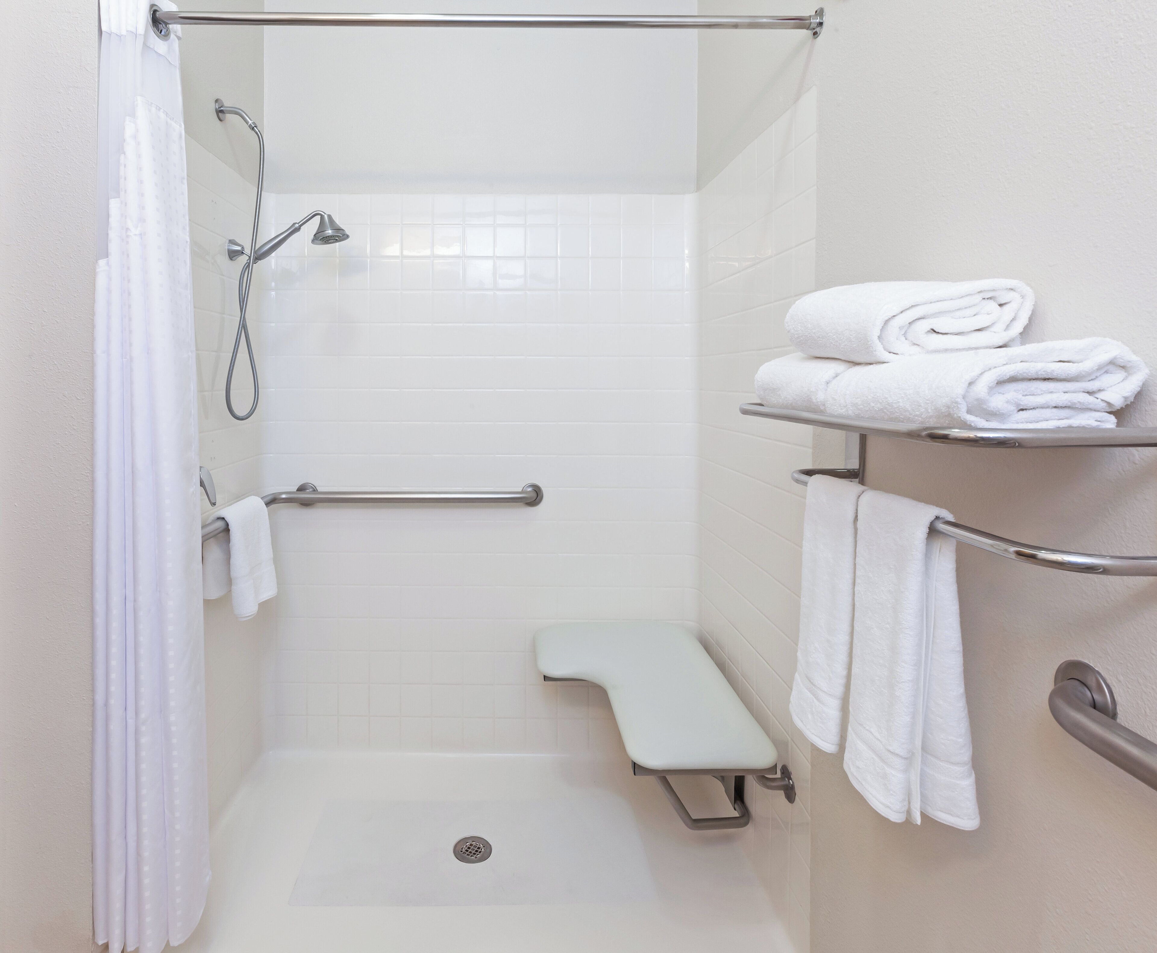 Un espace douche équipé d’un pommeau de douche à main, d’une barre d’appui, d’un banc et d’un porte-serviettes avec des serviettes pliées.