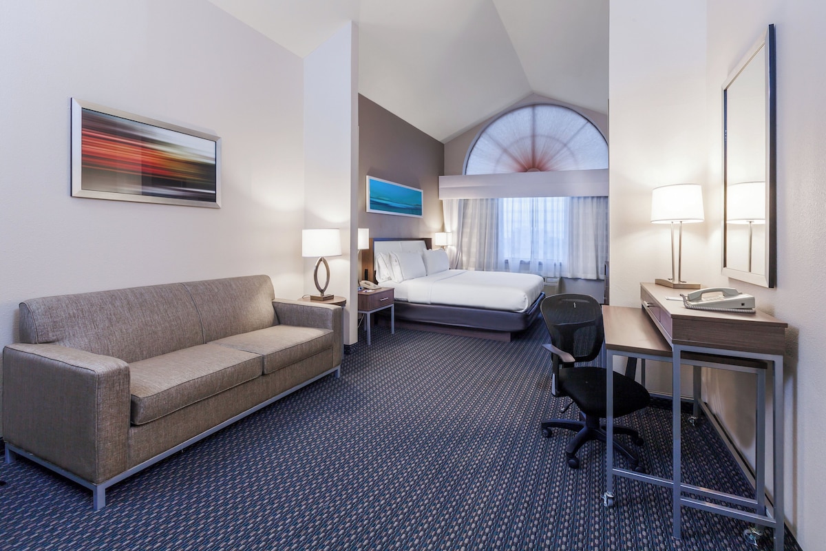 Ein Hotelzimmer mit einer Couch, einem Bett, einem Schreibtisch mit Stuhl, einer Schreibtischlampe, einem Telefon und einem Bild an der Wand.