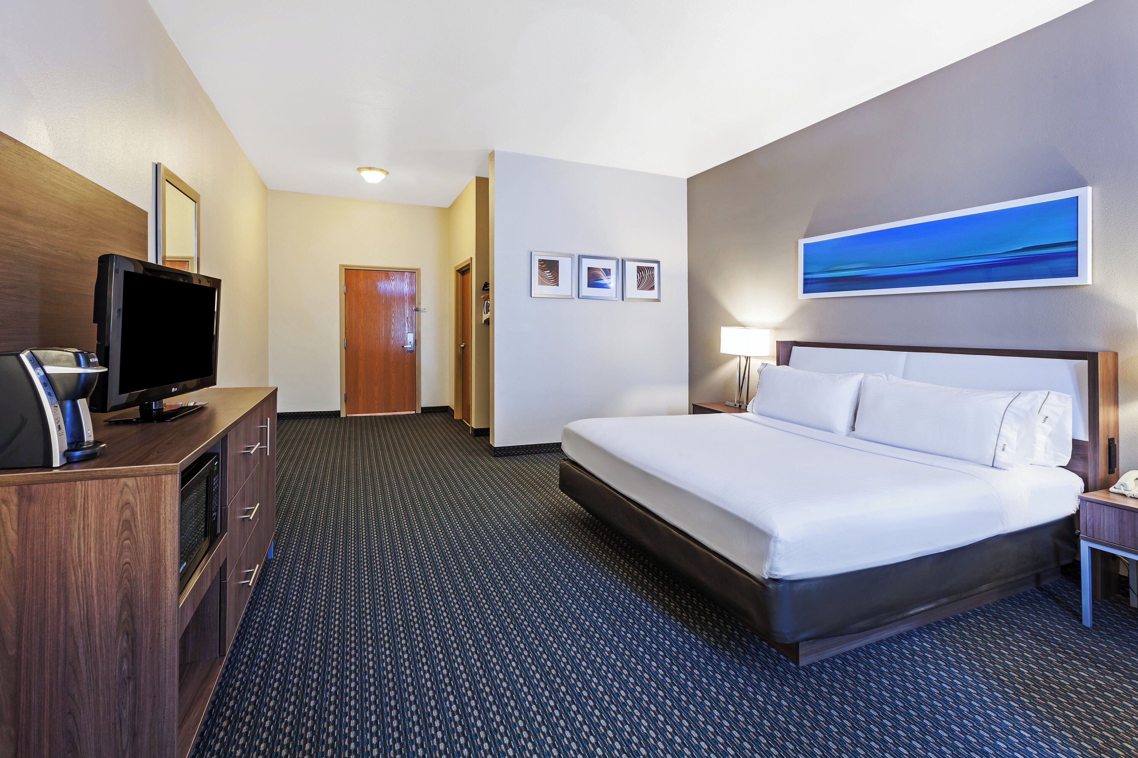 A hotel room with a large bed, a desk with a flat-screen TV, a coffee machine, and a door leading to another room.