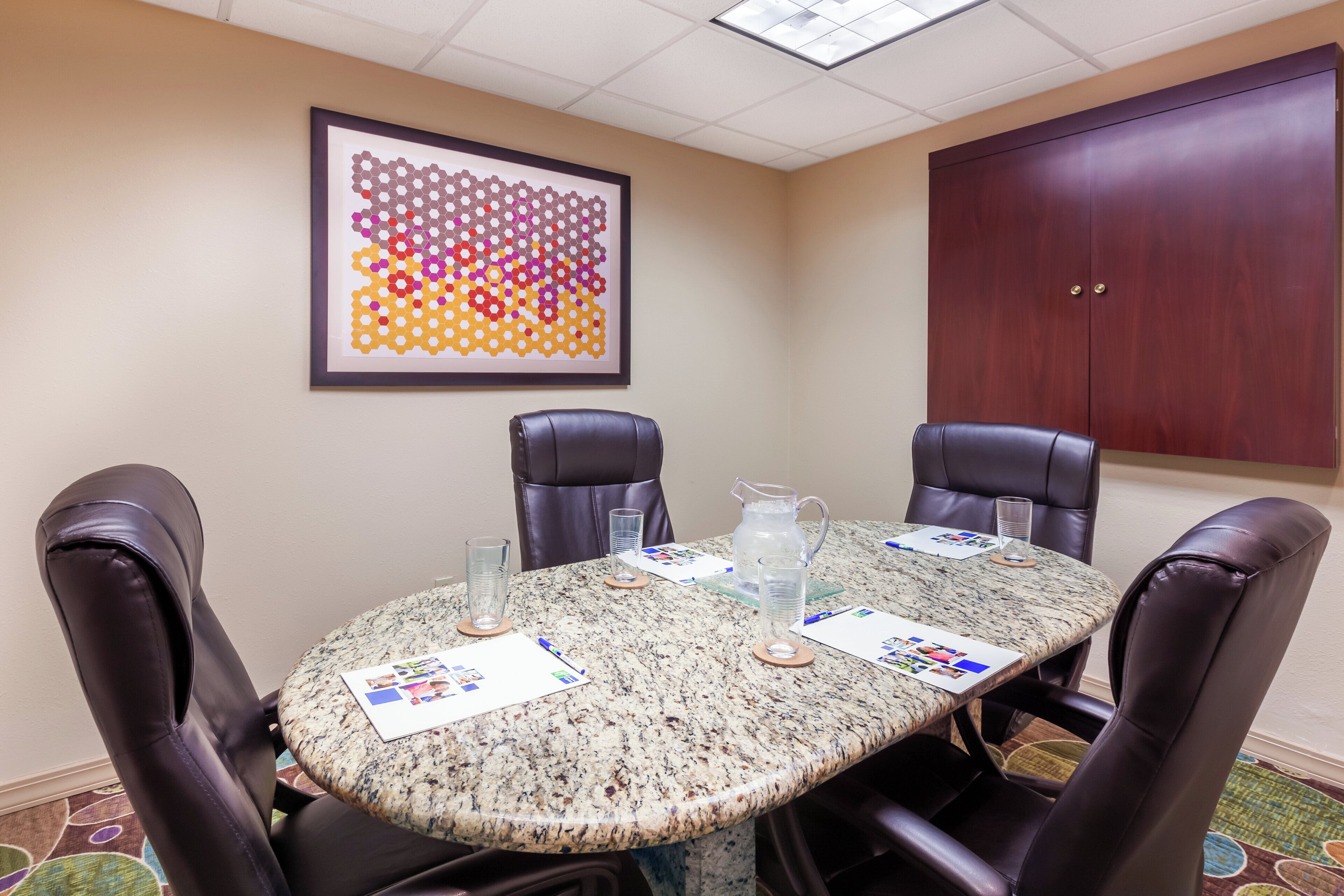 A conference room with a round table, four leather chairs, a framed abstract art piece, a wooden cabinet, and a water pitcher.