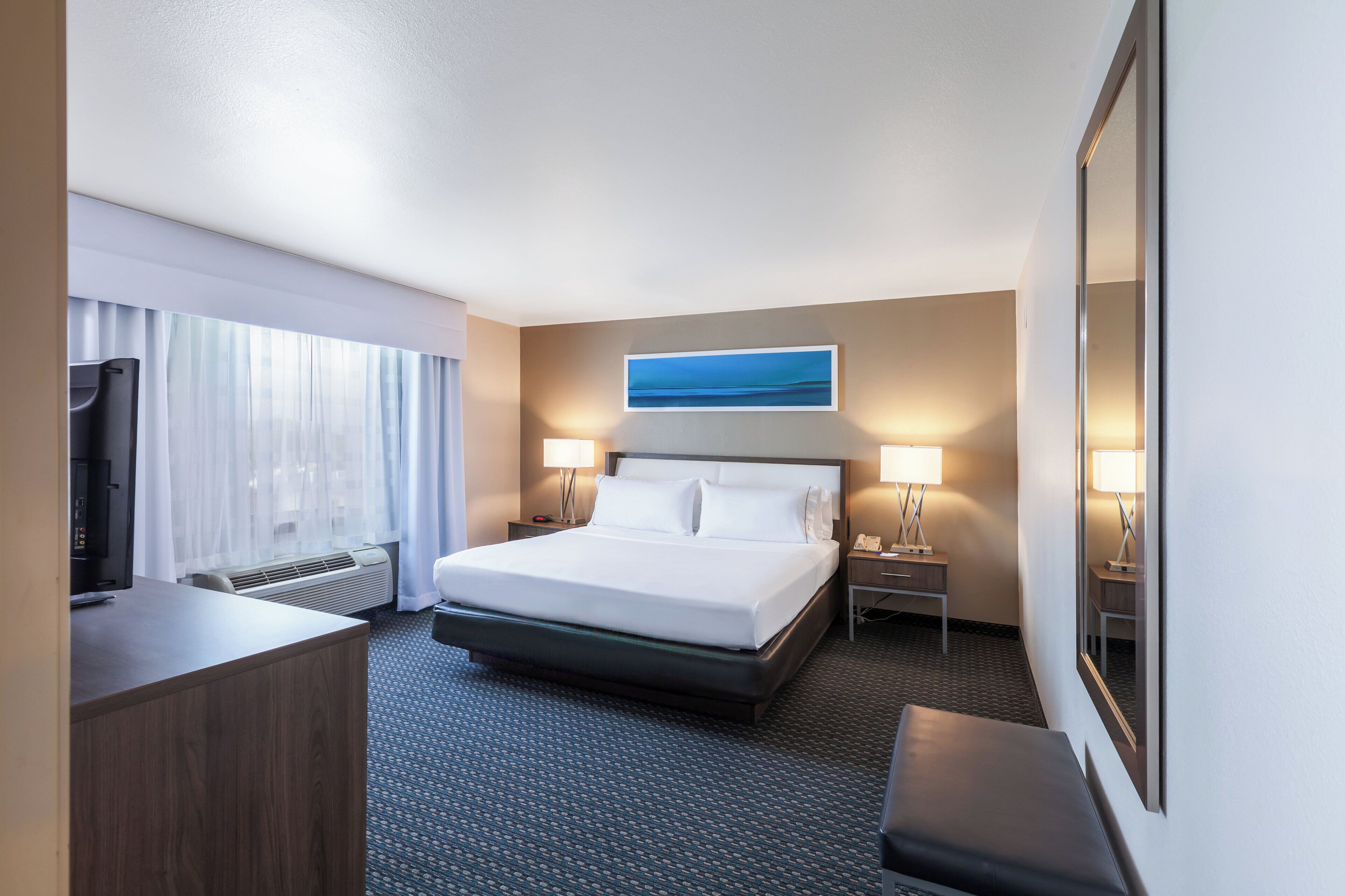 Una habitación de hotel con cama, lámparas de noche, escritorio con televisor y un gran ventanal con cortinas.