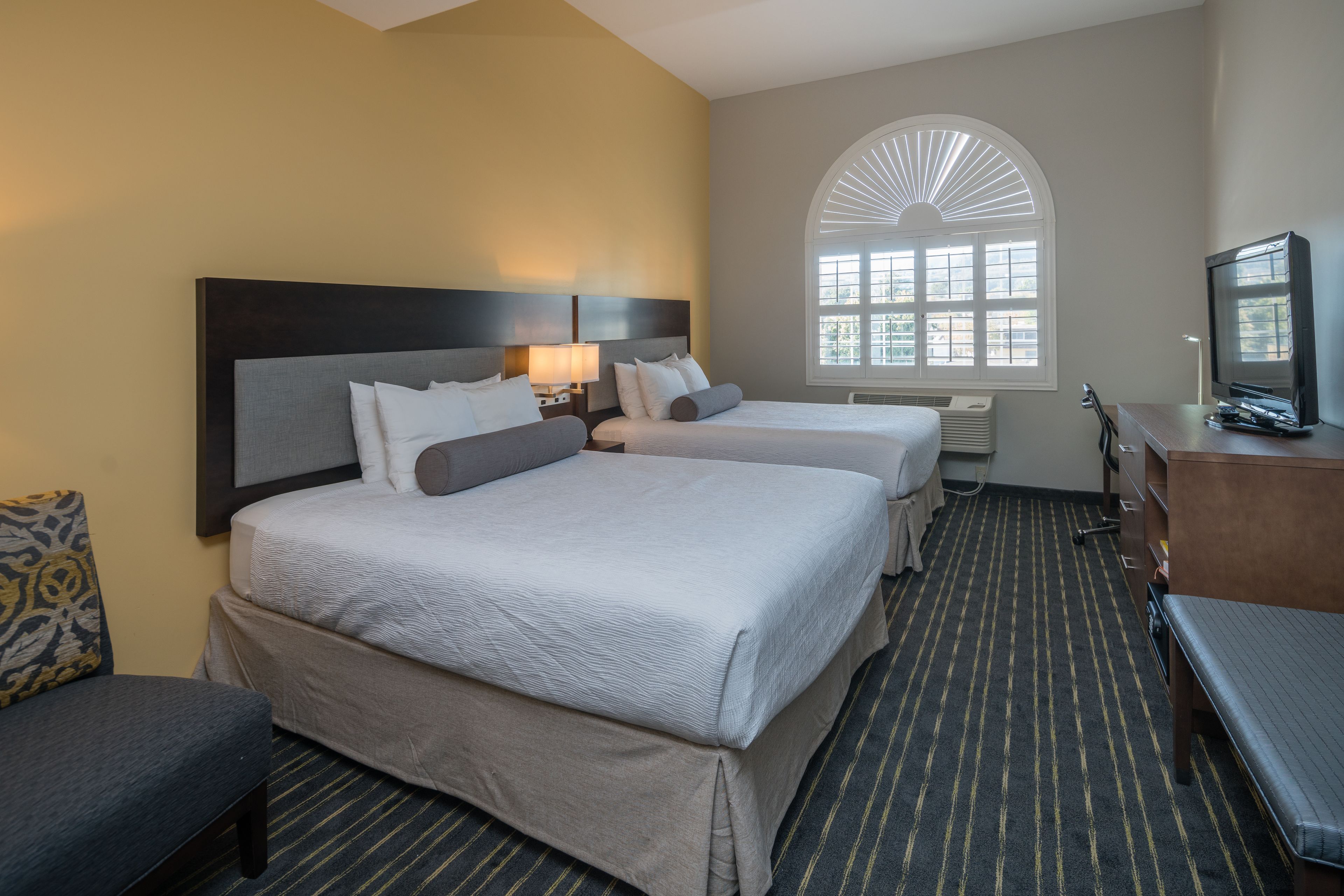 A hotel room with two beds, a desk, a television, and a large arched window.