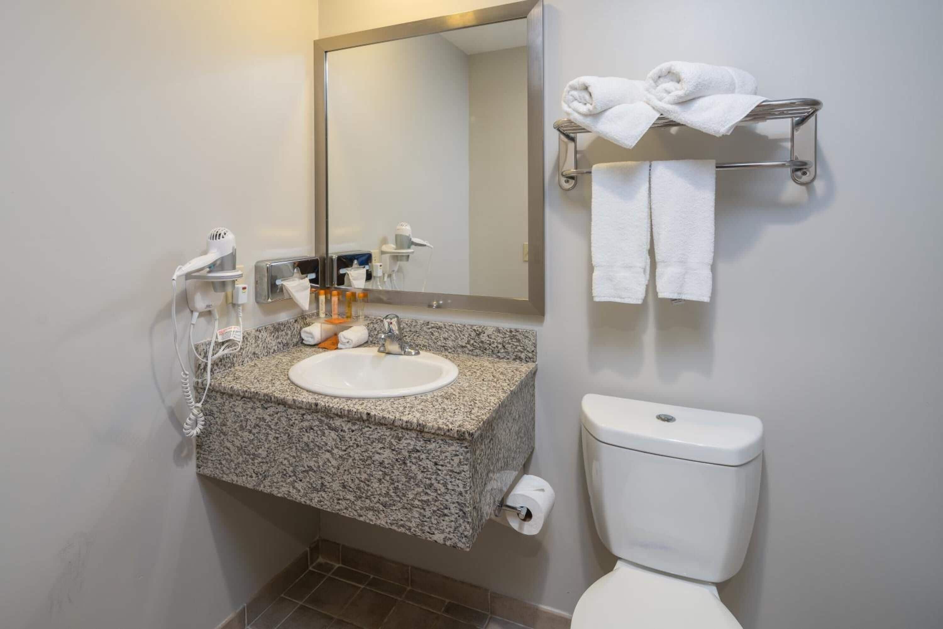 A bathroom with a granite countertop sink, a mirror, a towel rack with towels, a toilet, and a hairdryer.
