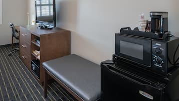 A hotel room with a desk, a computer, a microwave, a coffee maker, and a bench.
