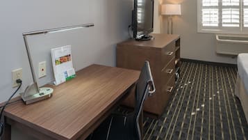 Hotellihuone, jossa on puinen työpöytä, televisio, joka on sijoitettu kaappiin, lamppu, tuoli ja sänky.