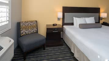 A hotel room with a bed, a chair, a nightstand, a lamp, and a heater.