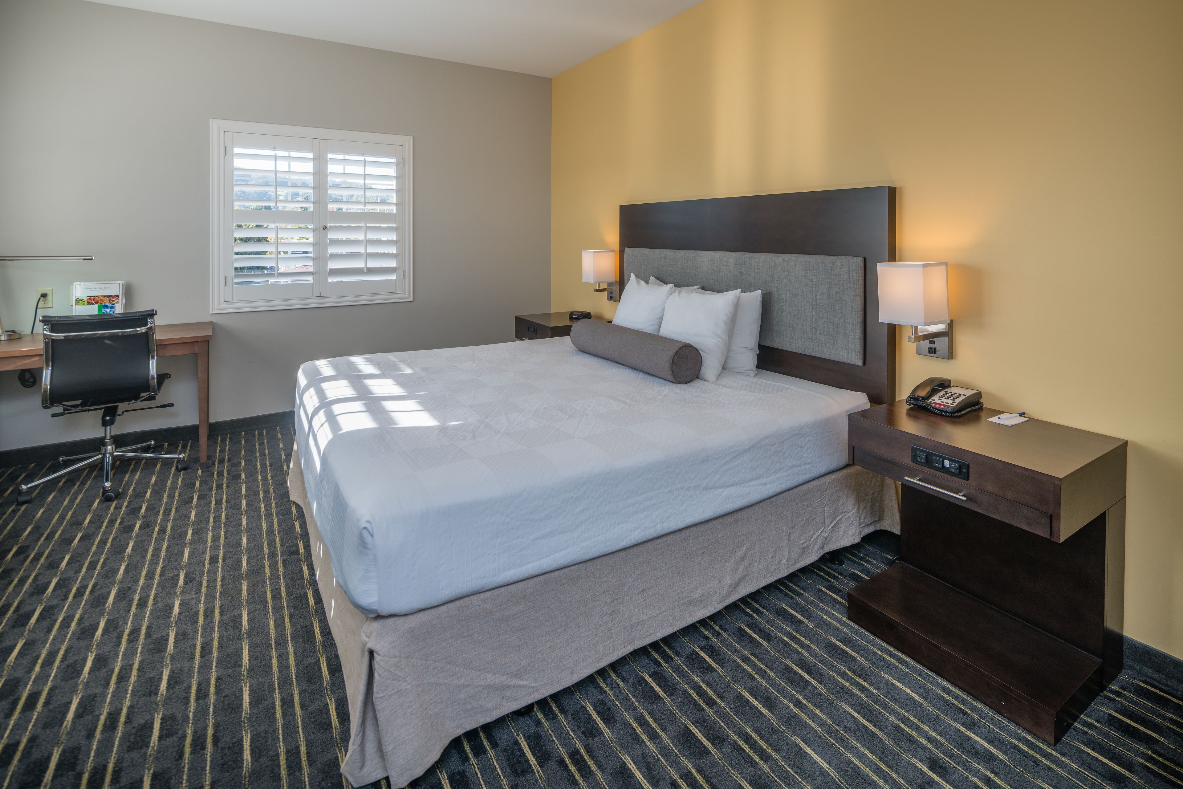 A hotel room with a large bed, a desk, a chair, a window with blinds, and a nightstand with a lamp.