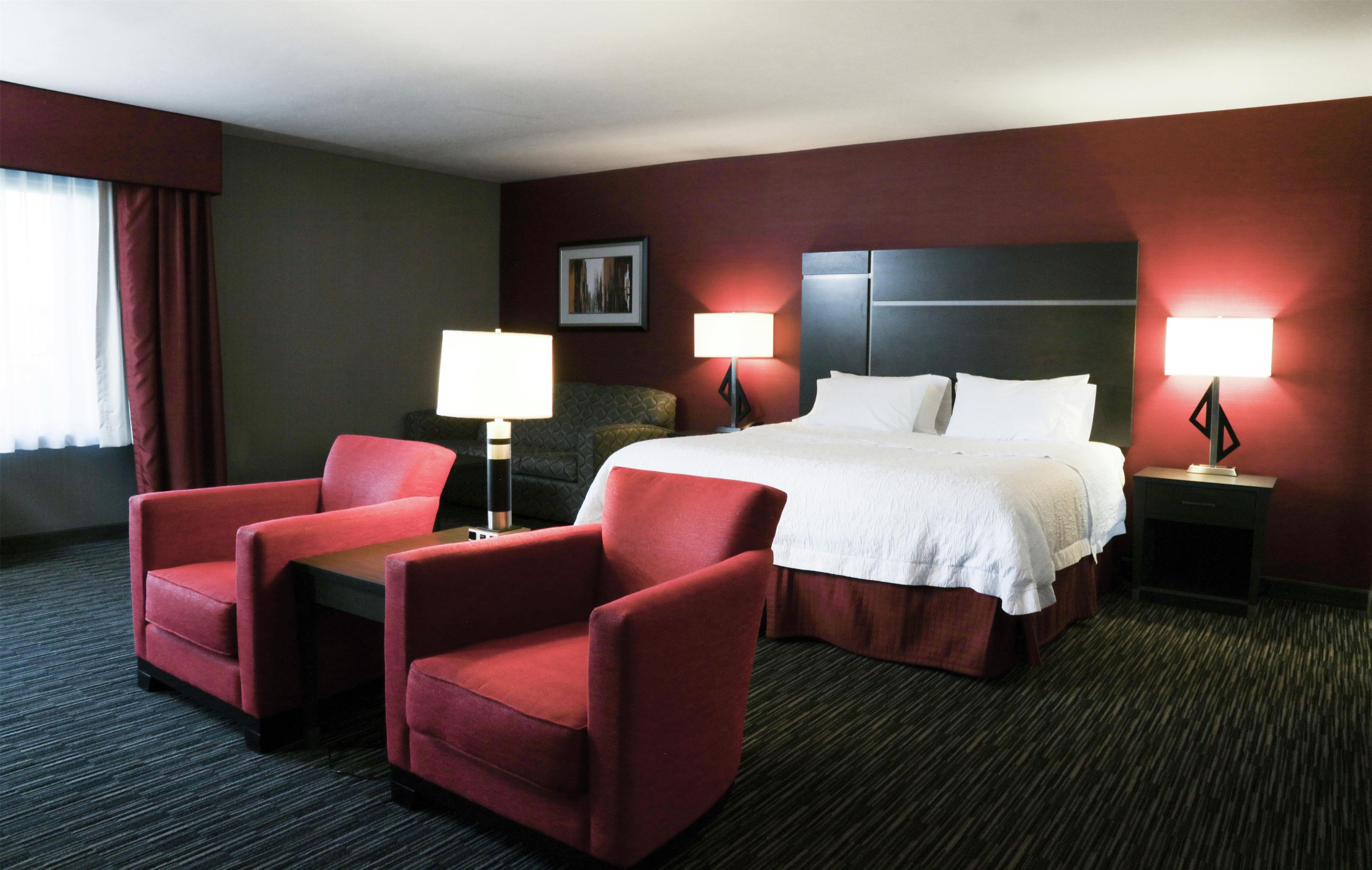 Une chambre d’hôtel avec un lit, deux fauteuils rouges, deux tables de chevet avec des lampes, un tableau encadré au mur et une fenêtre avec des rideaux.