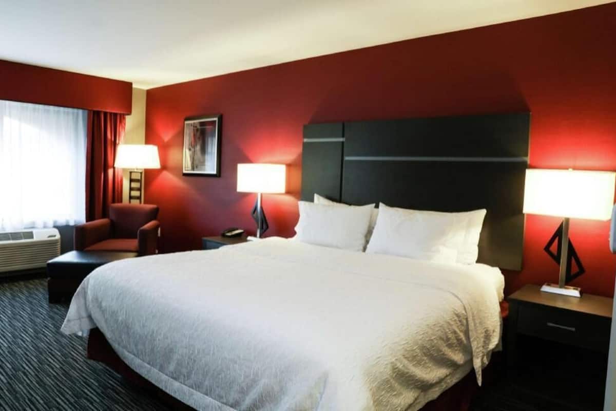 Ein Hotelzimmer mit einem großen Bett, zwei Nachttischen mit Lampen, einem Sessel und einem Fenster mit Vorhängen.