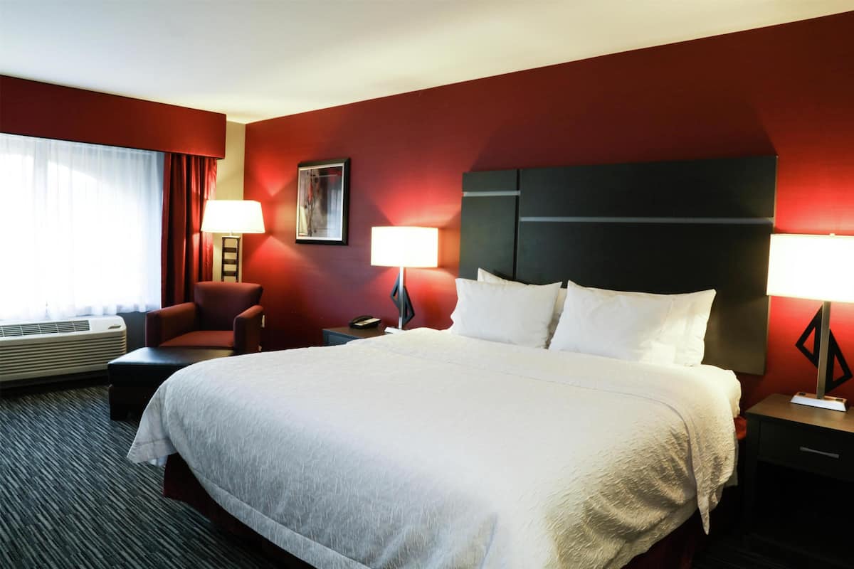 Ein Hotelzimmer mit einem großen Bett, zwei Nachttischen mit Lampen, einem Sessel, einem Gemälde an der Wand und einem Fenster mit Vorhängen.
