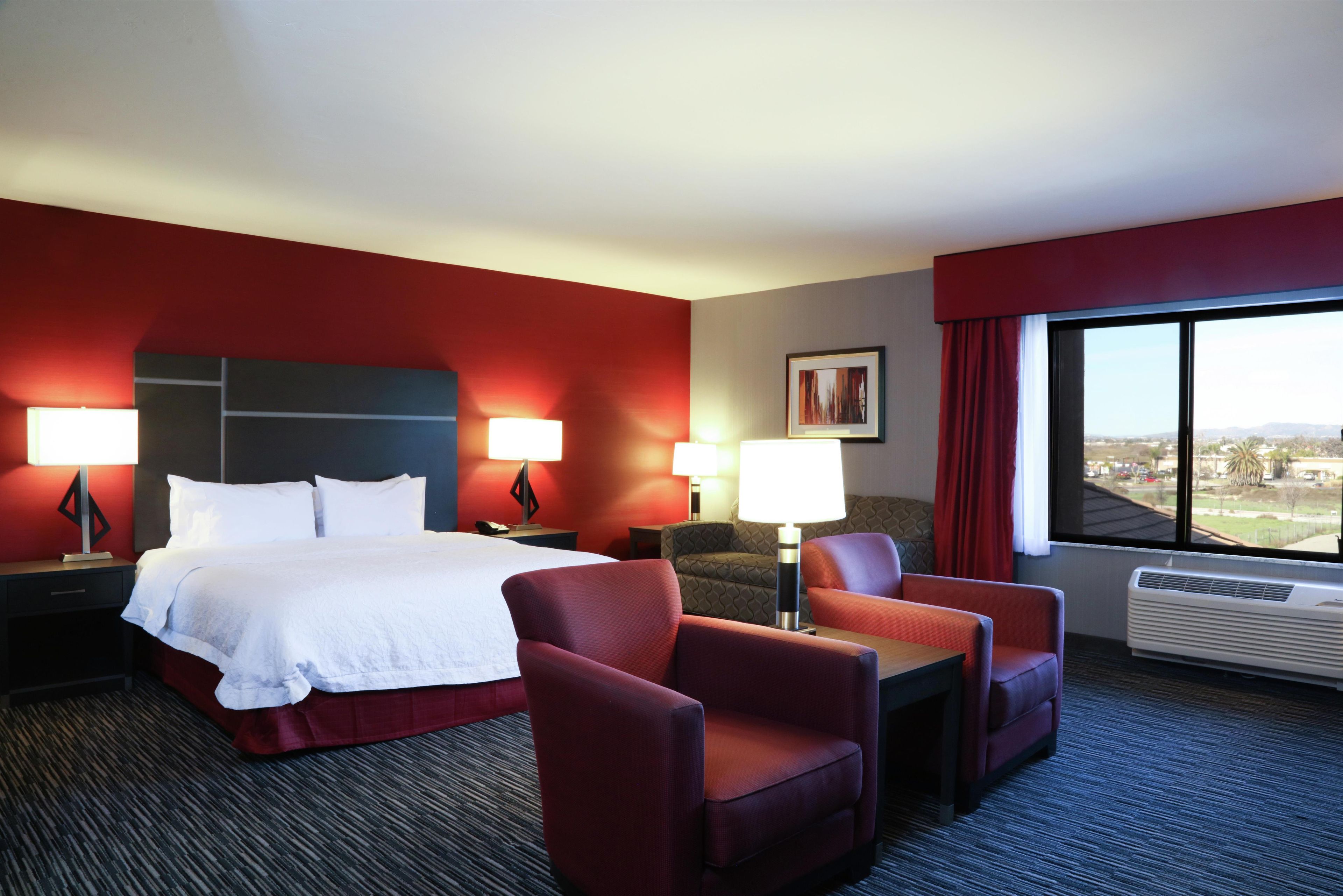 A hotel room with a bed, two armchairs, a desk, two bedside lamps, a framed picture on the wall, and a large window with a view.