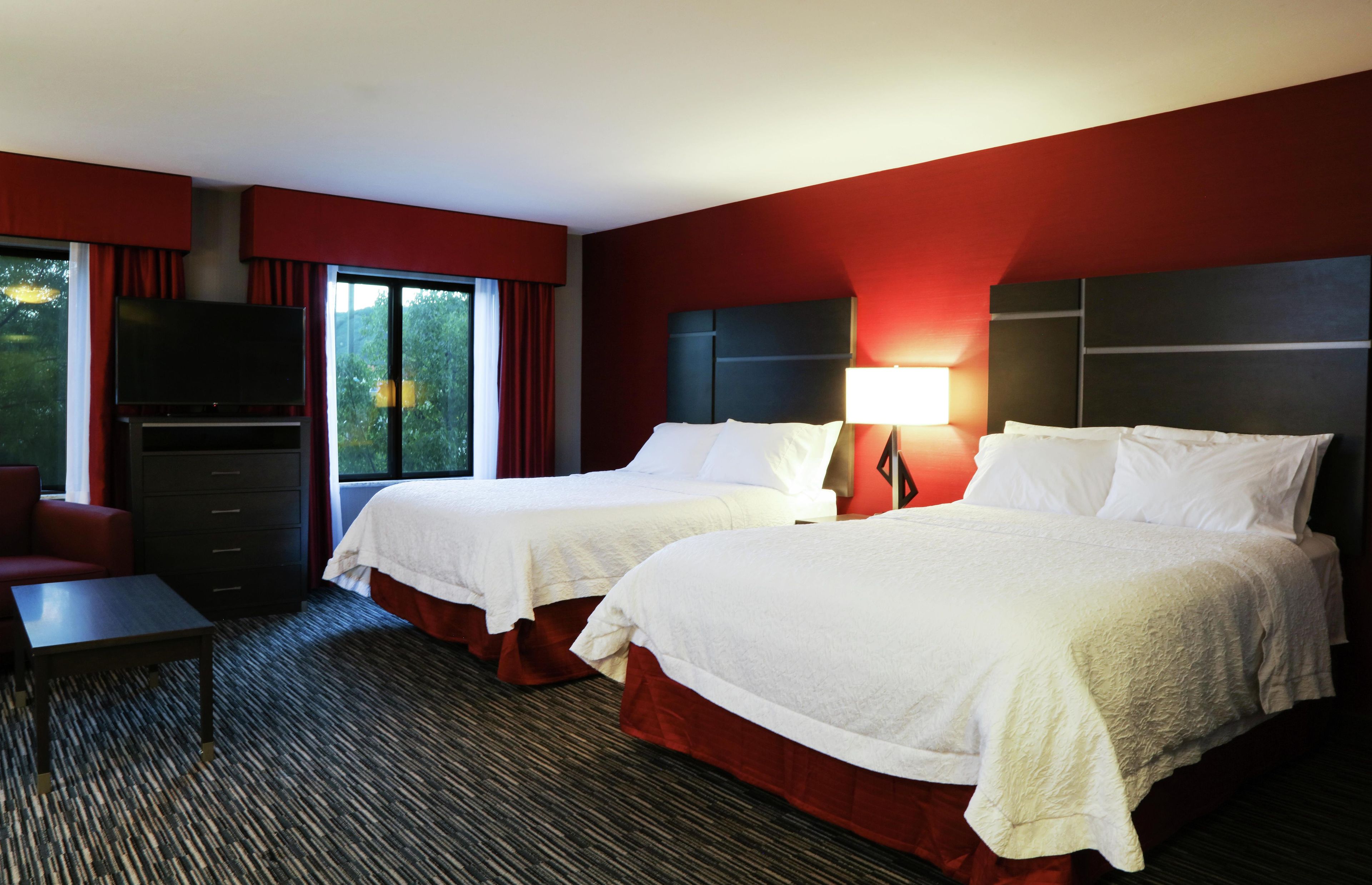 A hotel room with two beds, a TV, a desk, and a window with curtains.