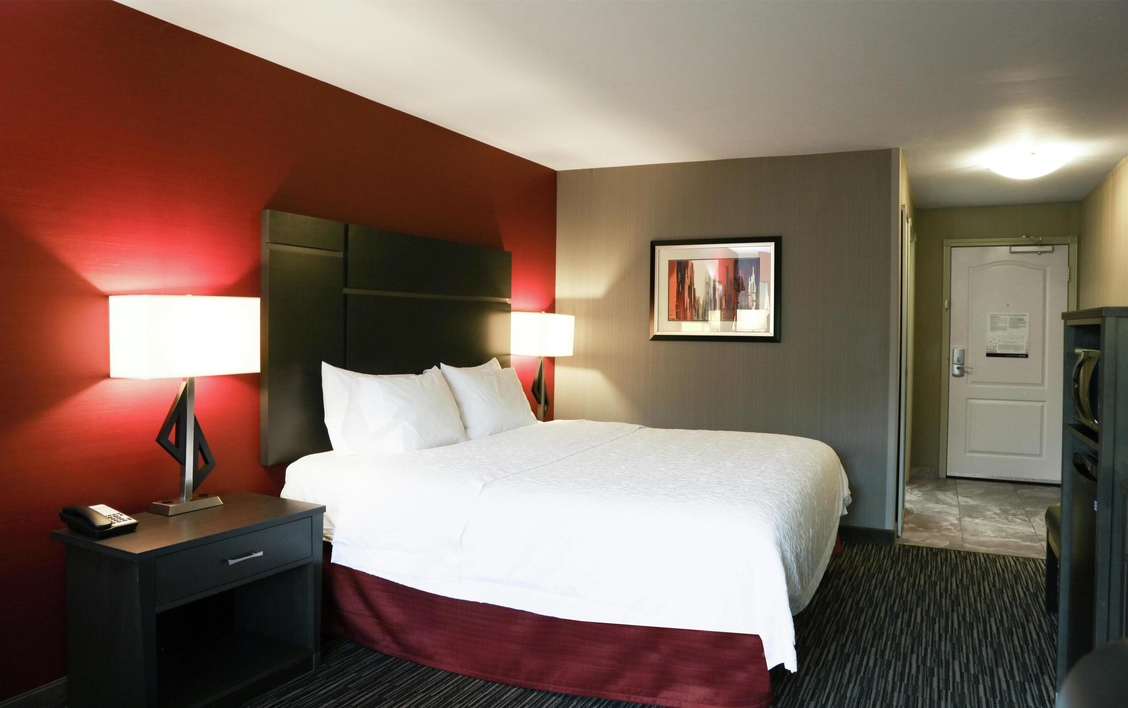 A hotel room with a bed, bedside lamps, a desk with a phone, a door, and a framed picture on the wall.