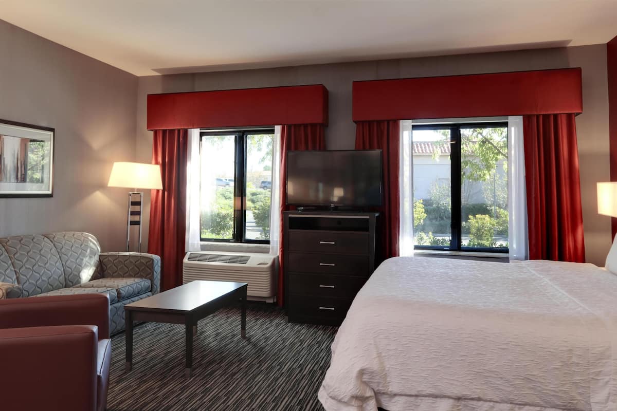 Ein Hotelzimmer mit Bett, einer Couch, einem Fernseher, einem Schreibtisch und einem Fenster mit Vorhängen.