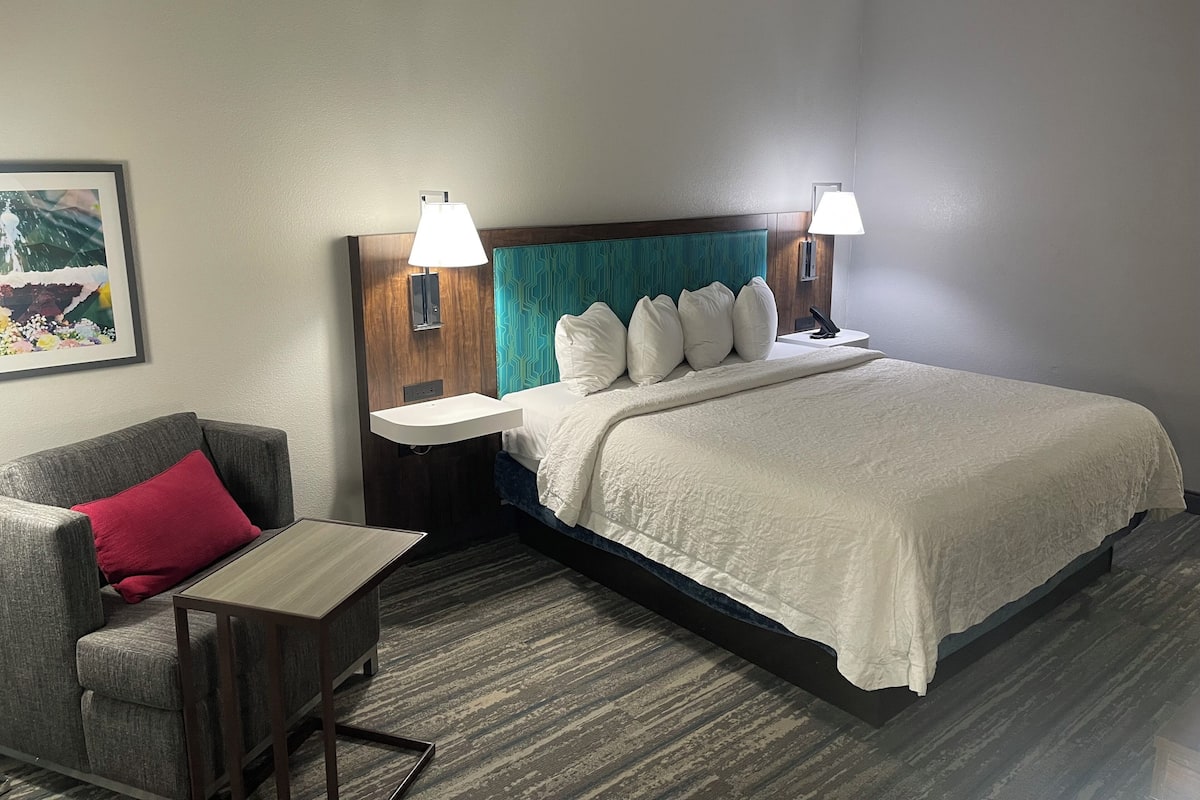 Ein Hotelzimmer mit Bett, Nachttisch, Stuhl, Lampe und einem Gemälde an der Wand.