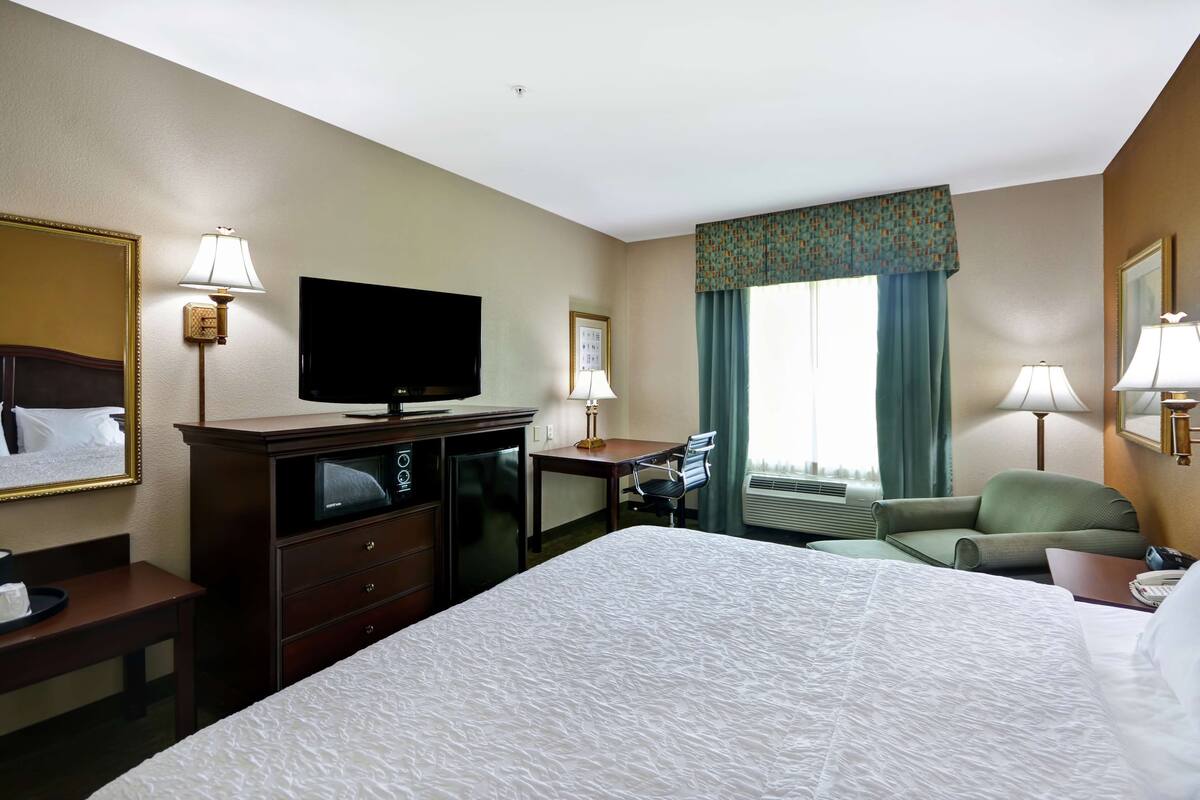 Ein Hotelzimmer mit einem großen Bett, einem Schreibtisch mit Stuhl, einem Fernseher, einer Schreibtischlampe und einem Fenster mit Vorhängen.