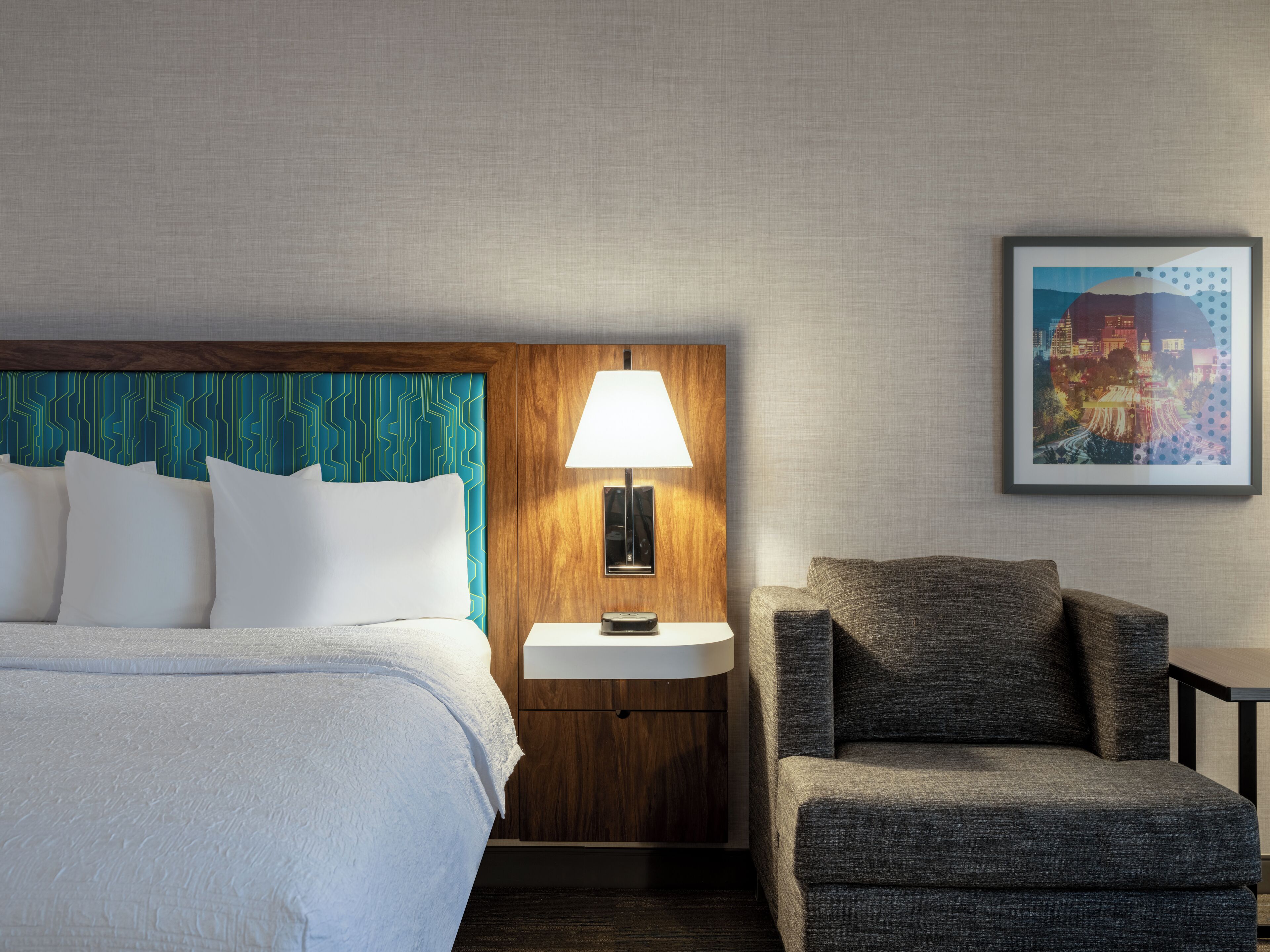 Habitación de hotel con cama, mesita de noche con lámpara, una silla e imagen enmarcada en la pared.