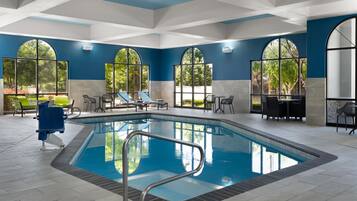 An indoor swimming pool with clear blue water, surrounded by lounge chairs and tables, large arched windows, and a tiled floor.