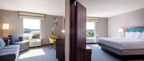 A modern hotel room with a large bed, a sofa, a desk, and a chair. There are large windows with views of the outside.