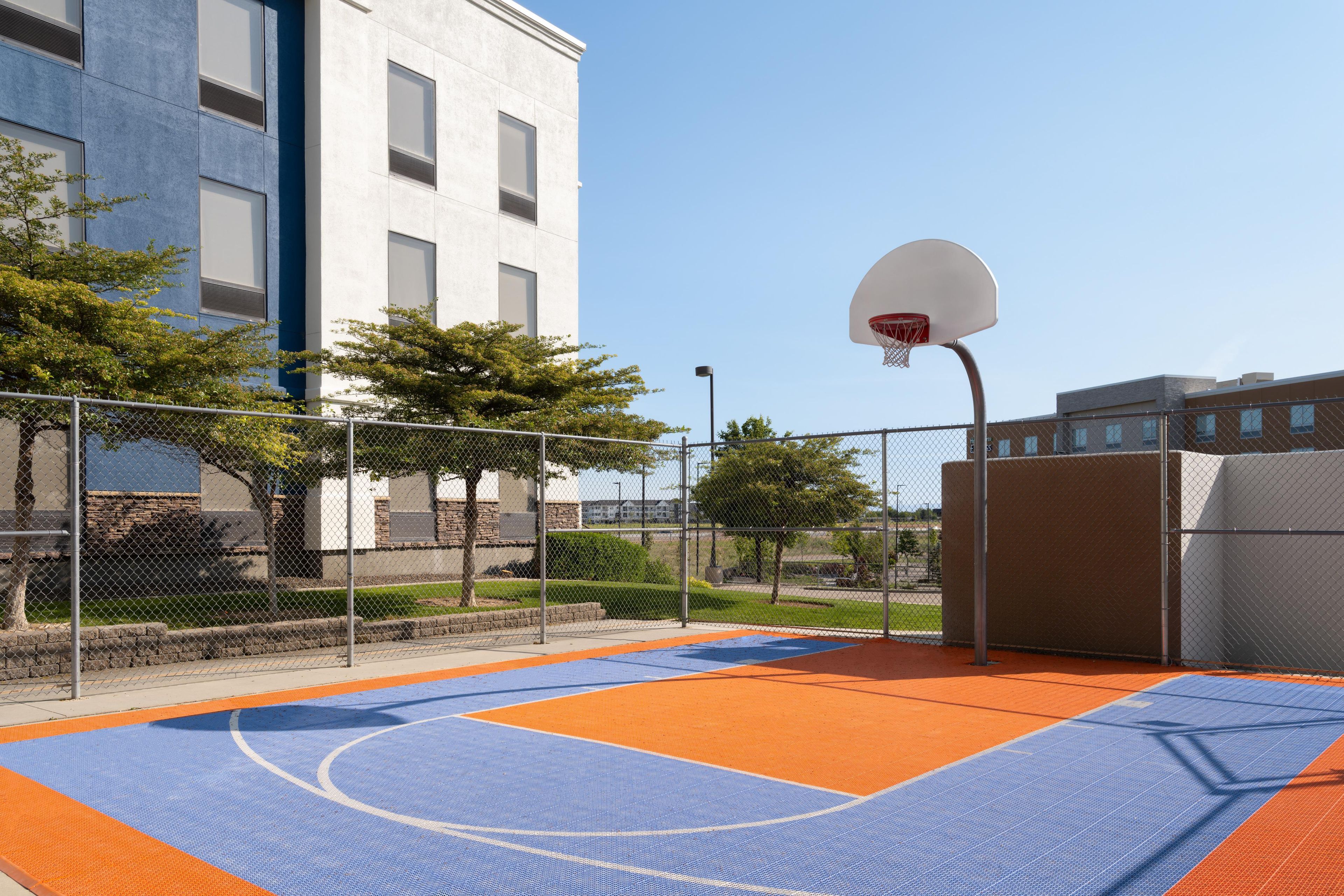 Un terrain de basket en plein air, entouré d’une clôture en treillis métallique, avec des arbres et un bâtiment en arrière-plan.