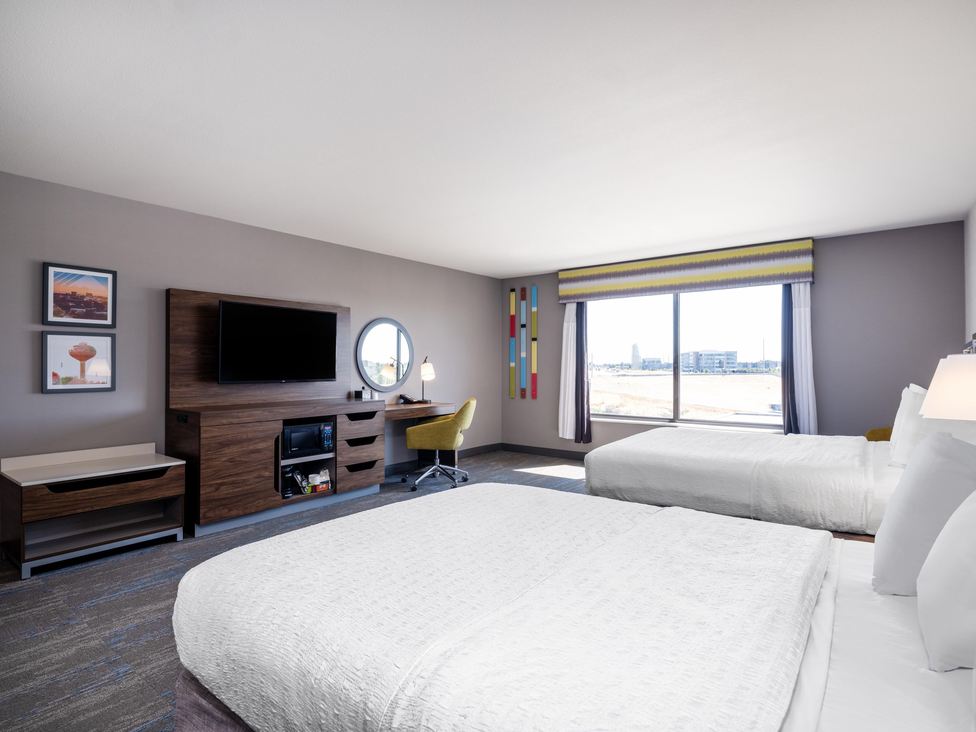 A hotel room with a large bed, a desk with a chair, a television, a mirror, and a window with a view of the city.