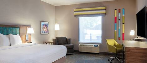 A hotel room with a large bed, a desk with a chair, a window with blinds, a wall-mounted TV, and a painting.