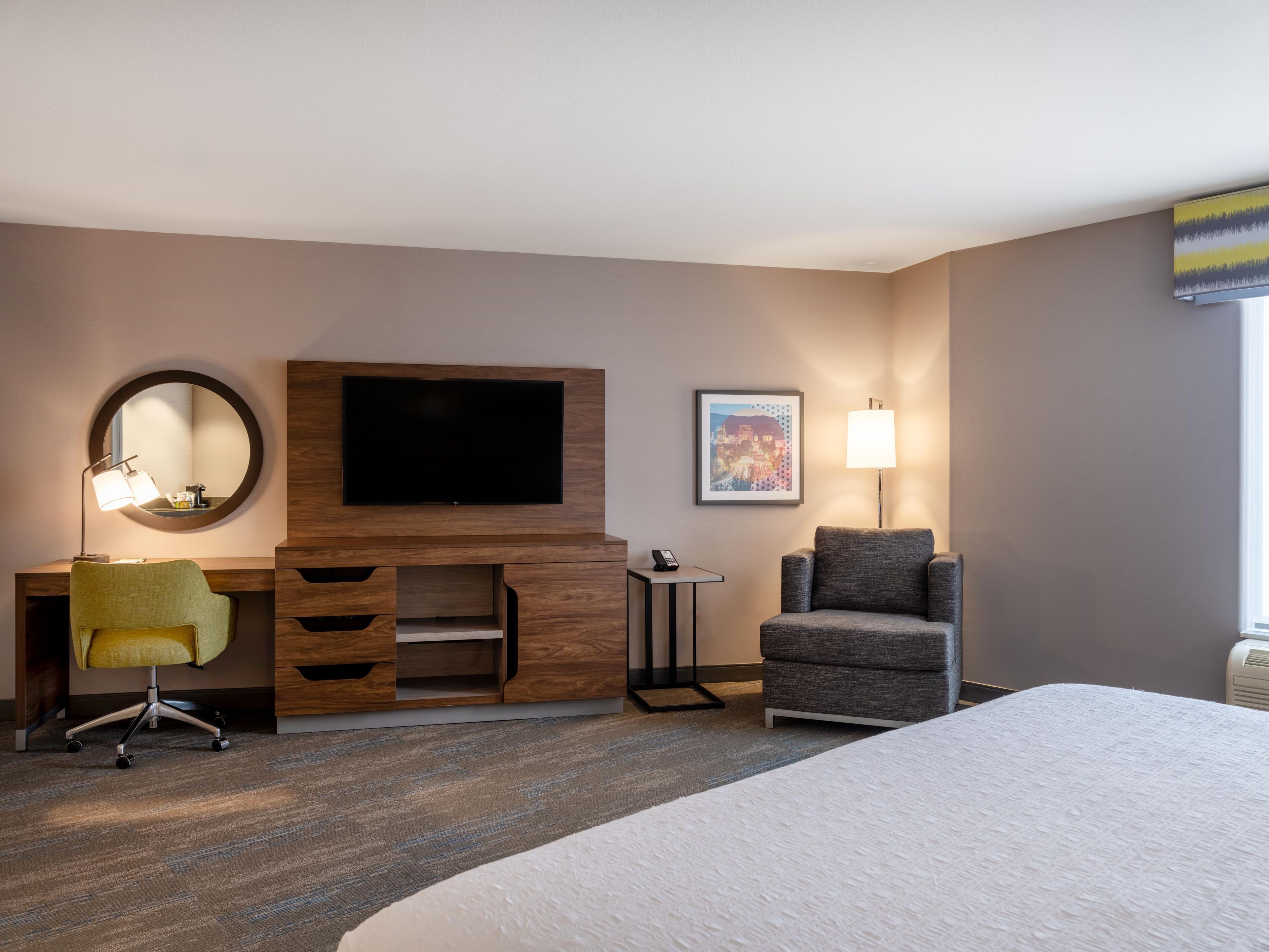 A hotel room with a bed, a desk with a flat-screen TV, a chair, a lamp, and a framed picture on the wall.