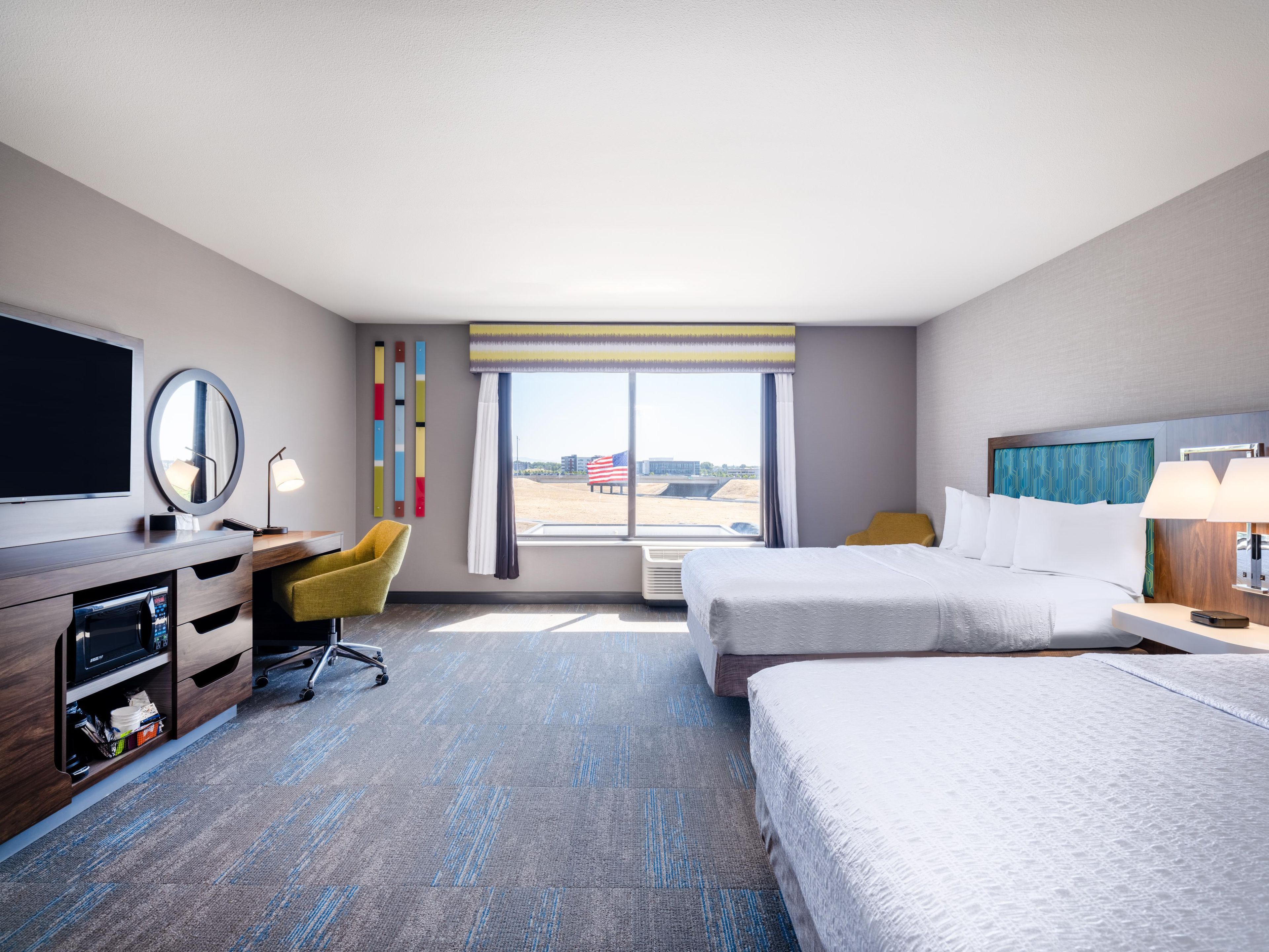 A hotel room with a bed, desk, chair, and a large window with a view of the airport.