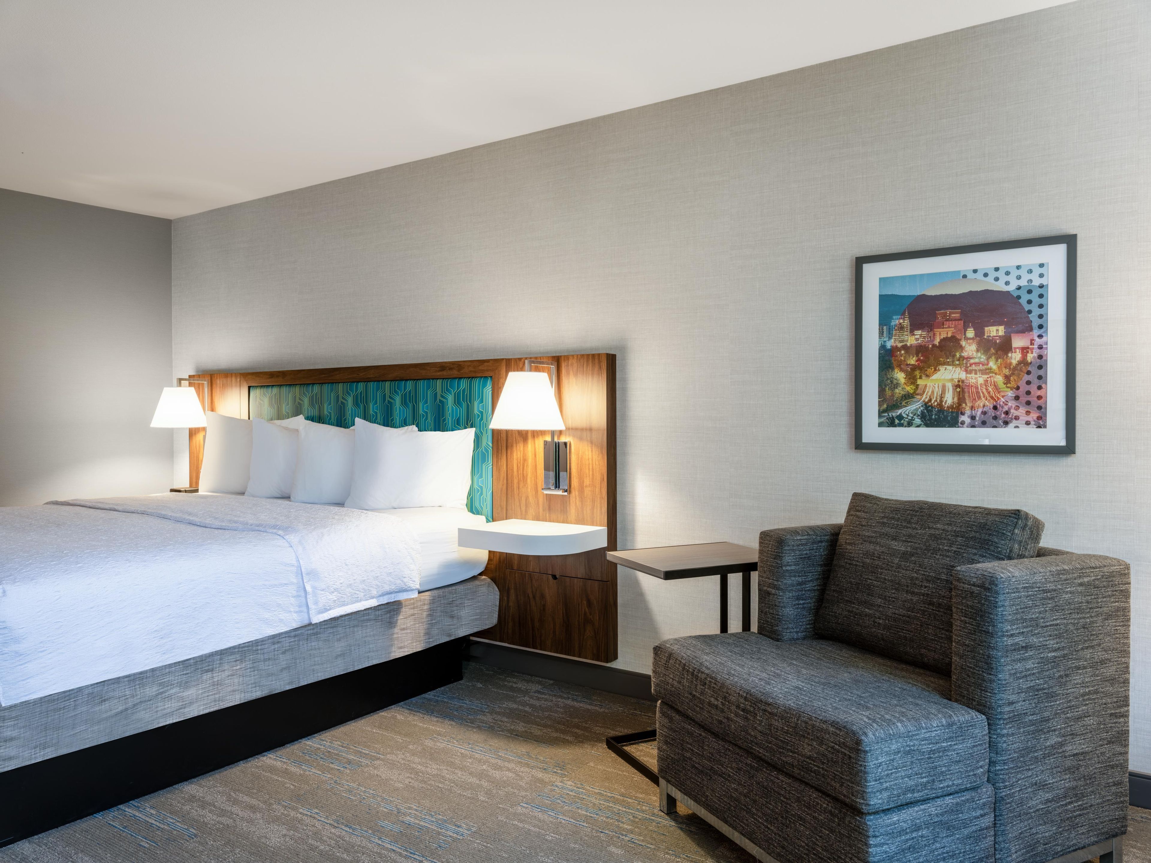 Habitación de hotel con cama, dos lámparas de noche, una mesita de noche, una silla y un cuadro en la pared.