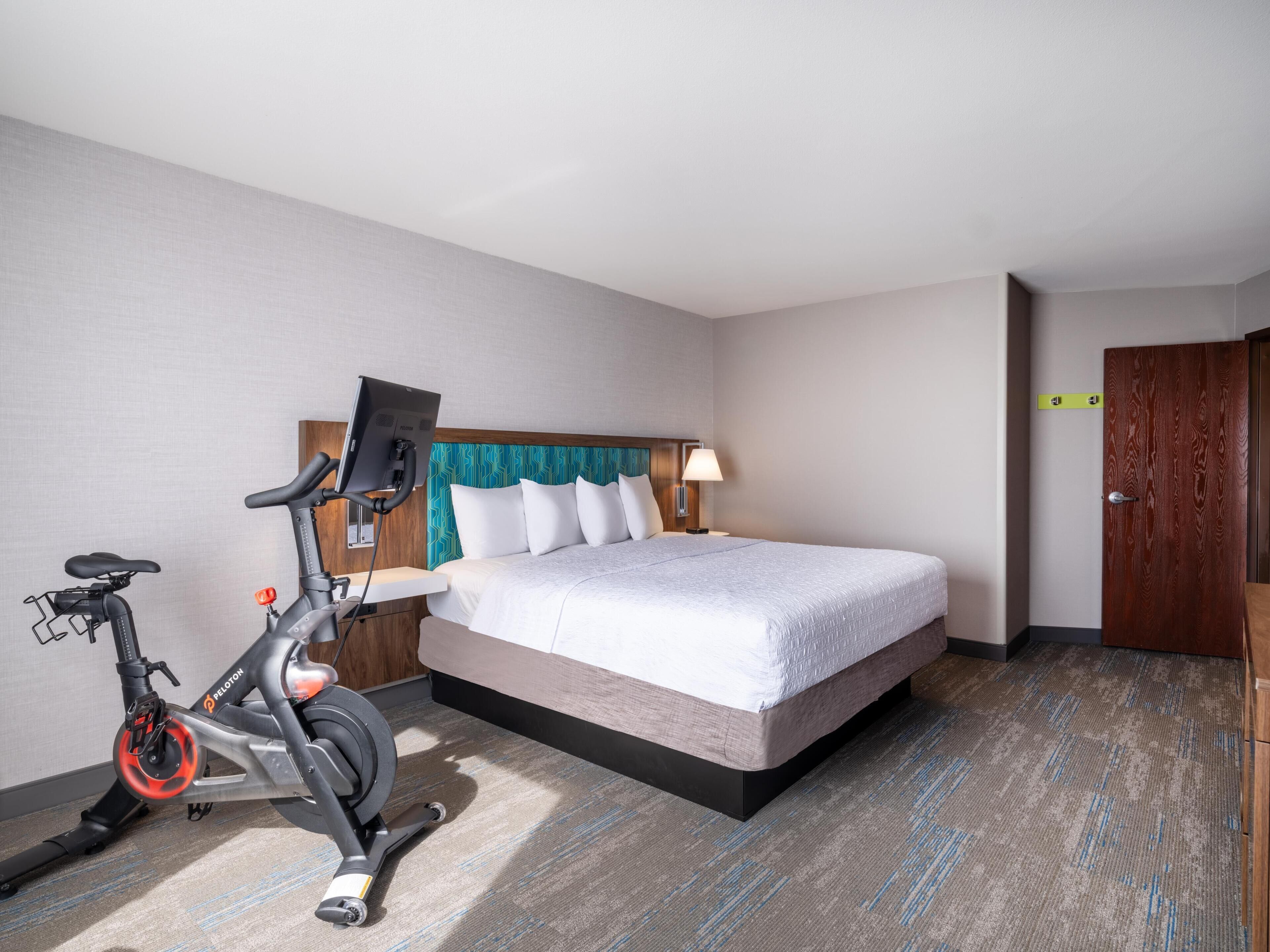Quarto de hotel com cama, secretária, cadeira, bicicleta ergométrica e porta.
