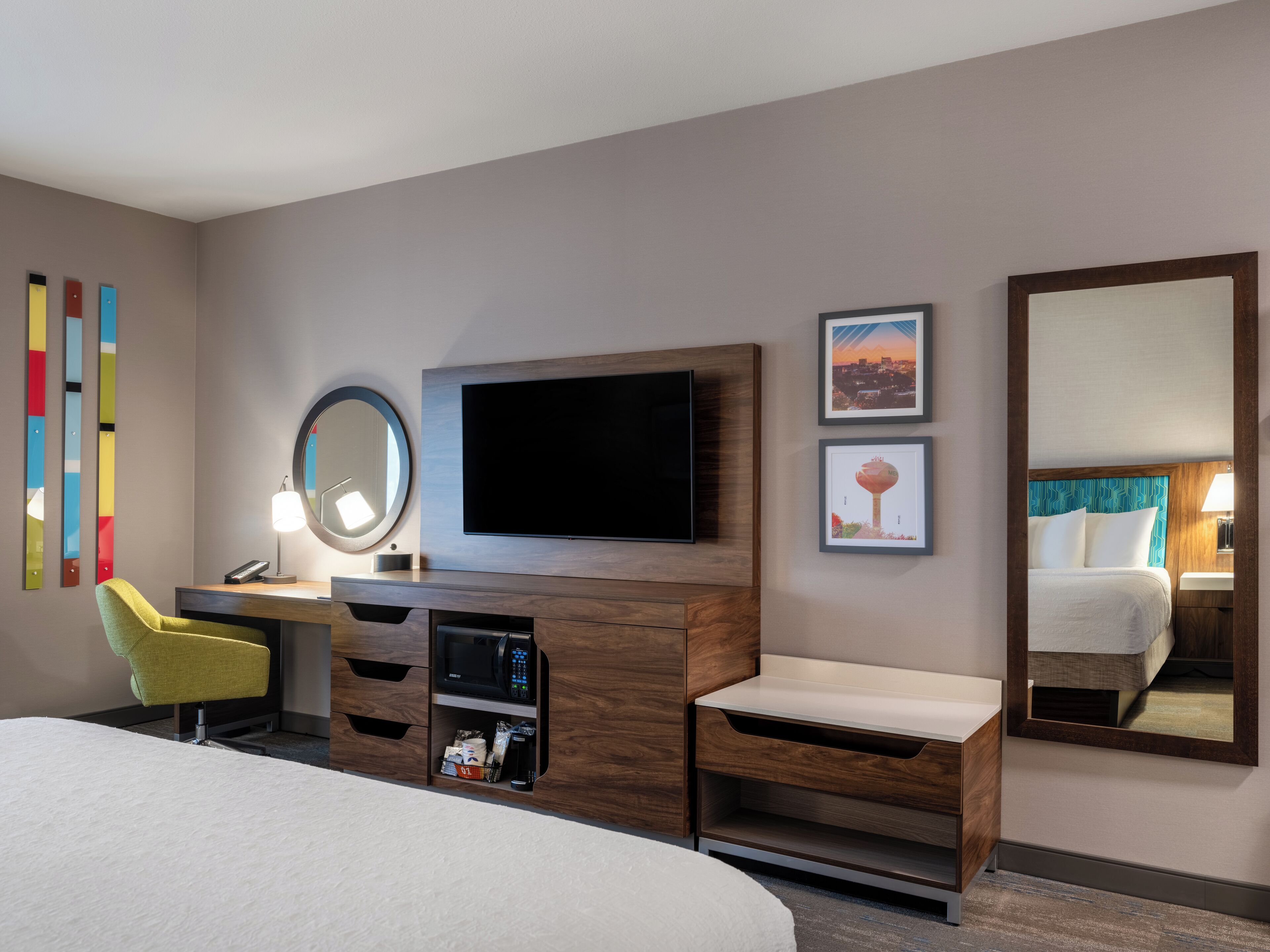 A hotel room with a bed, a desk with a chair, a TV, a mirror, and a dresser.