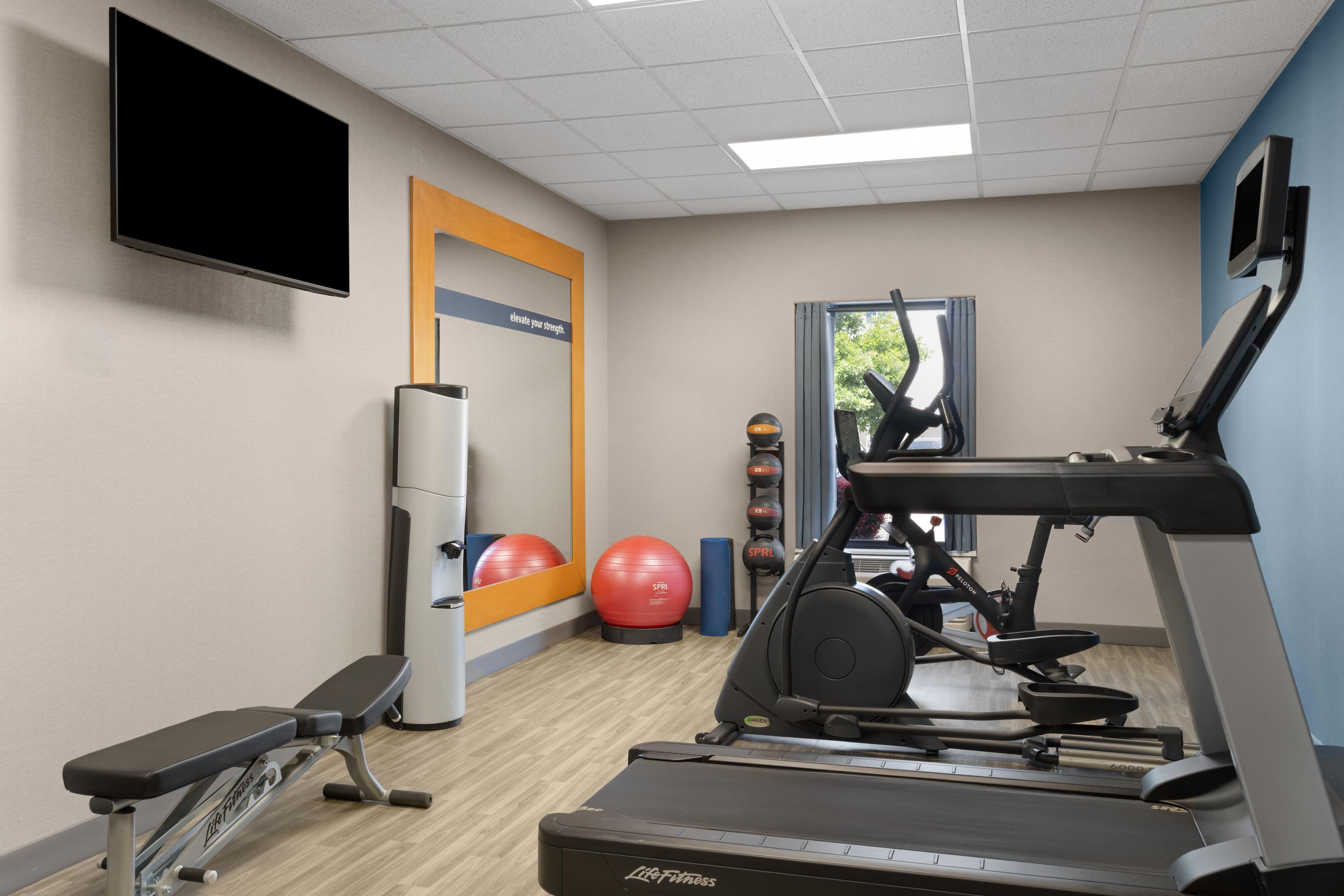 Academia com equipamentos de ginástica, um banco, uma TV fixada na parede e um cartaz motivacional.