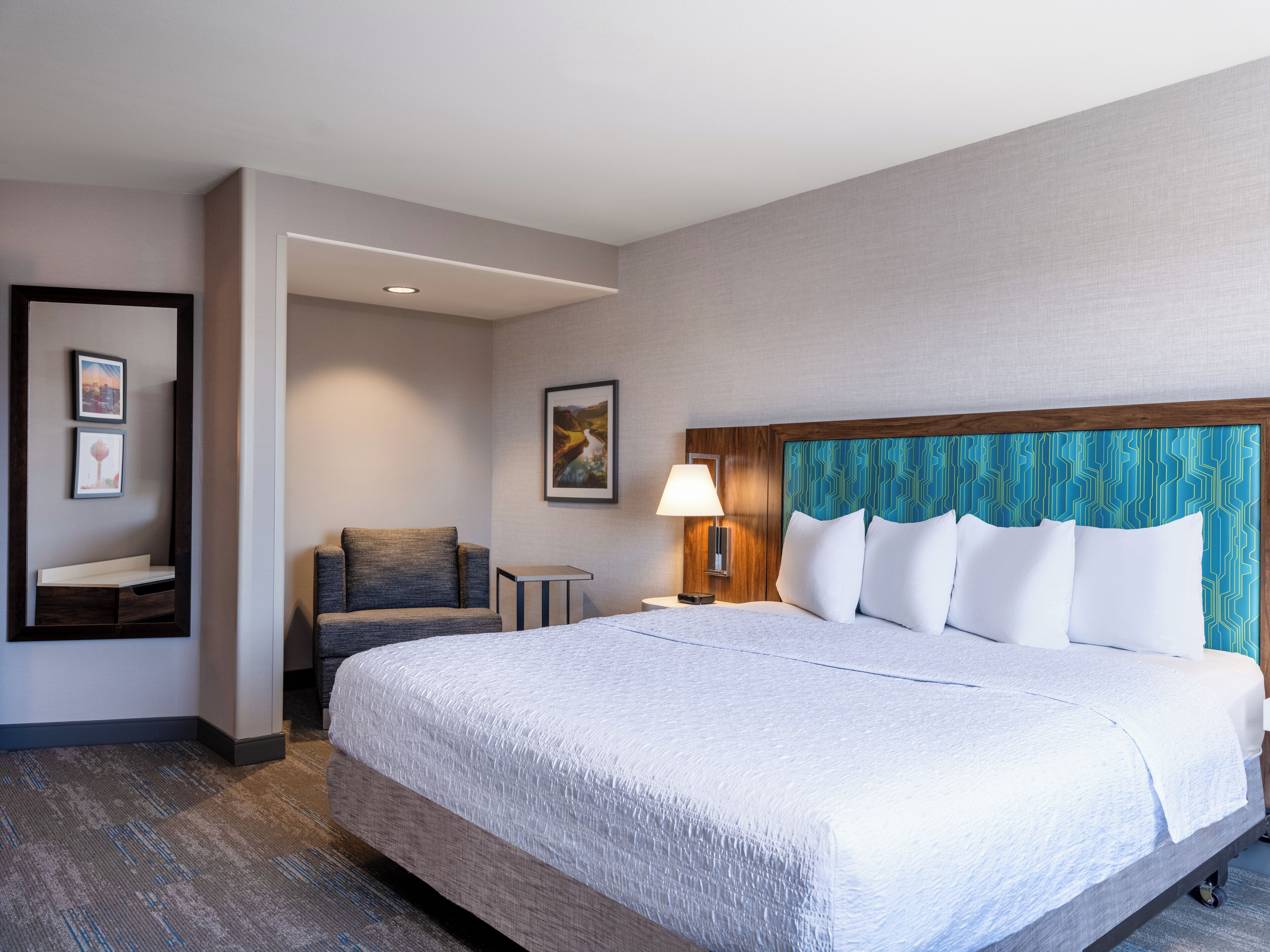 Habitación de hotel con una cama grande, un área de descanso con una silla y una mesita, y una lámpara de pared.