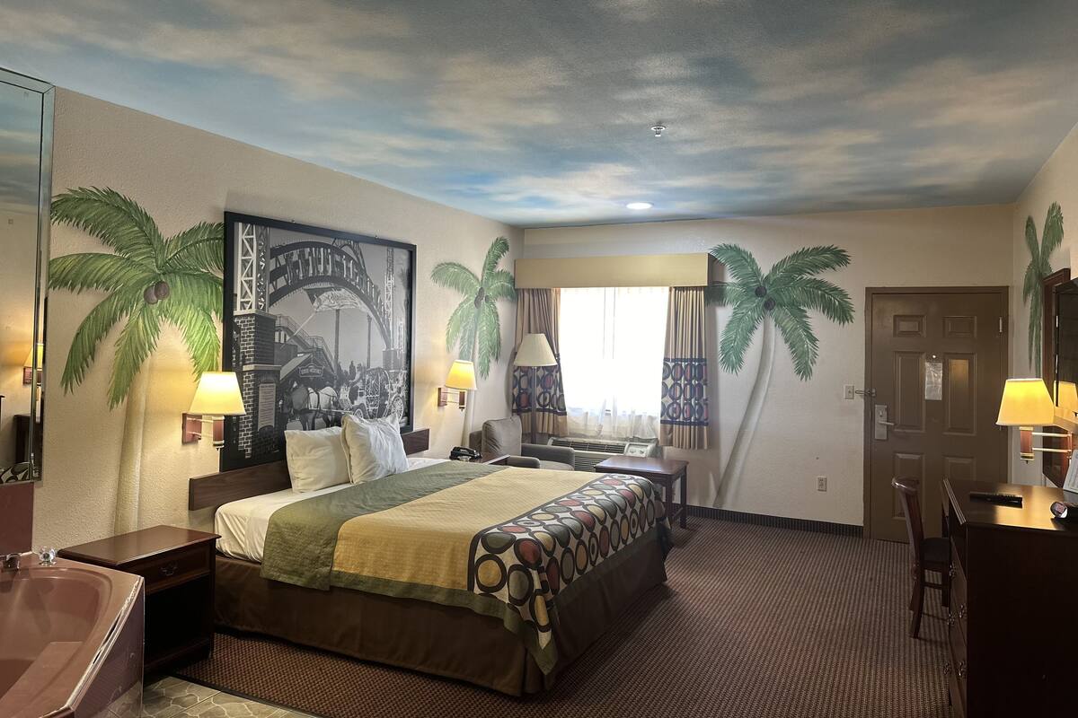 Une chambre d’hôtel comprenant un lit, un bureau, une chaise, une lampe et un grand tableau représentant un palmier et un pont.