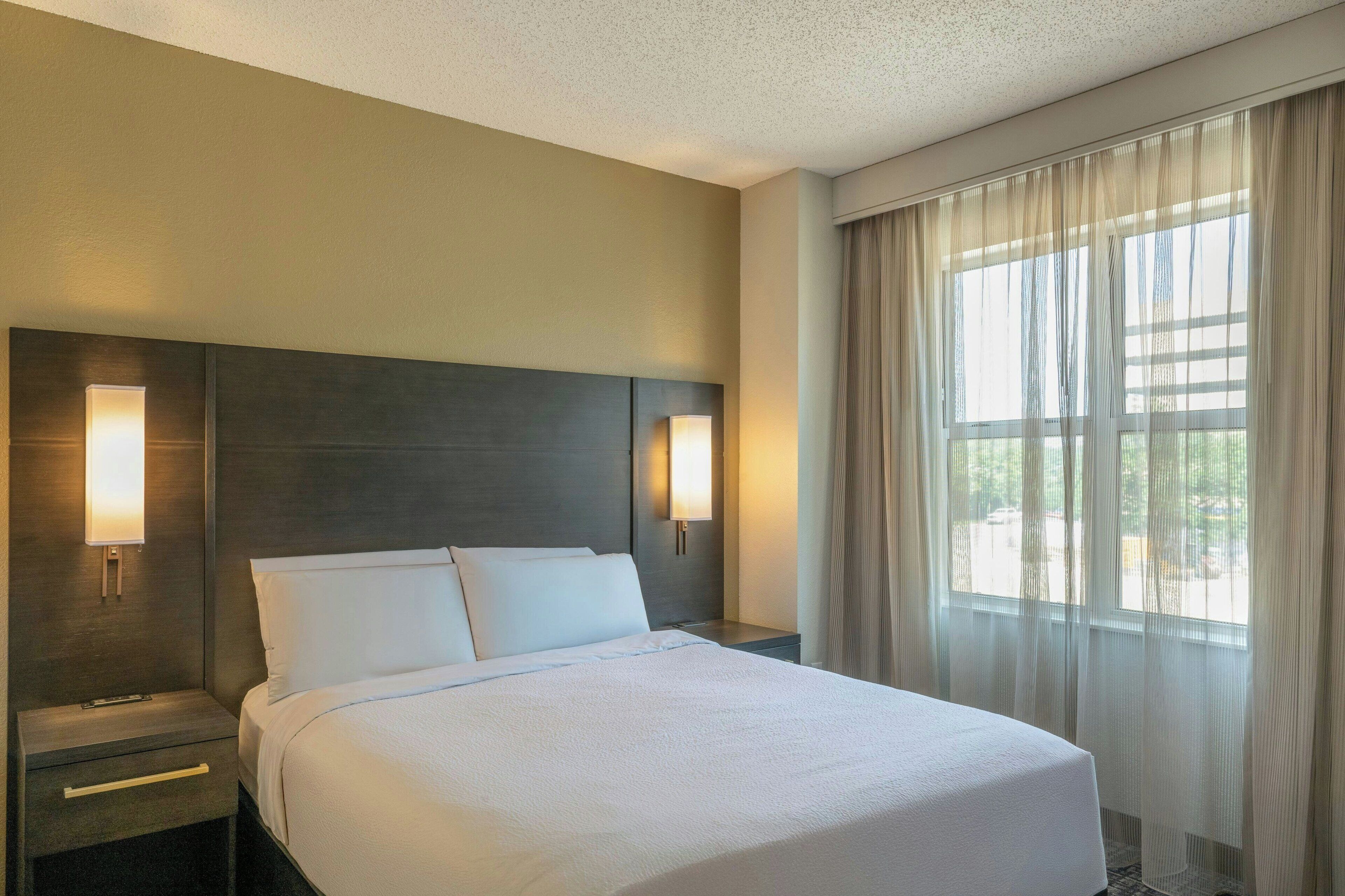A hotel room with a large bed, two bedside tables, and a window with sheer curtains.
