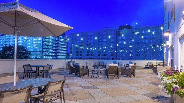 An outdoor terrace with tables and chairs, a large umbrella, and a view of a modern building with string lights. - Residence Inn by Marriott Rochester Mayo Clinic Area (Rochester)
