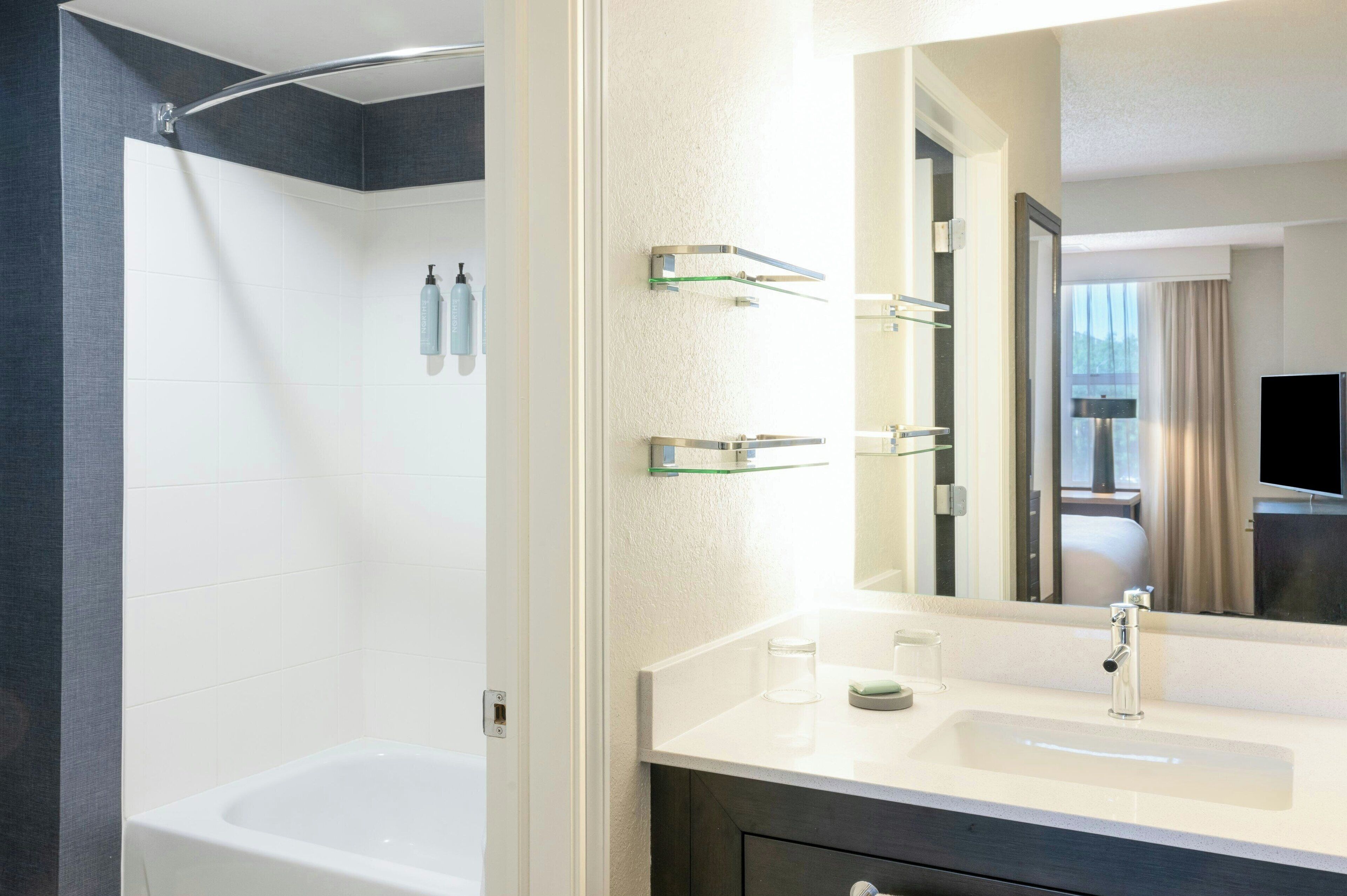 A modern bathroom with a bathtub, a large mirror, and a vanity with a sink and glass shelves.