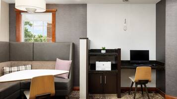 A modern hotel room with a gray sofa, a small dining table, two chairs, a kitchen area with a countertop and a TV, and a large window with a view of greenery.